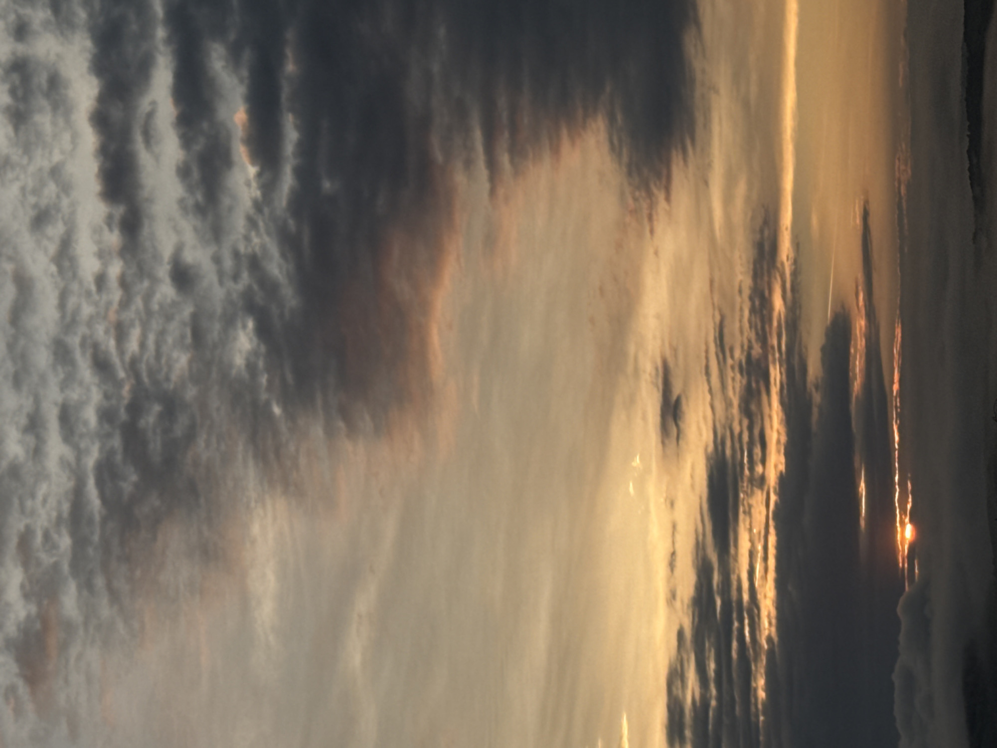 A vertical, eye-level, outdoor medium shot captures a dramatic sunset with a layer of fog or mist on the ground. The sky is filled with clouds, ranging from dark gray to light peach and orange. The sun, a bright orange orb, is partially visible through a gap in the clouds near the bottom of the frame, casting a warm glow. The fog below obscures the details of the landscape, but suggests a hilly terrain. The overall mood is serene and awe-inspiring.