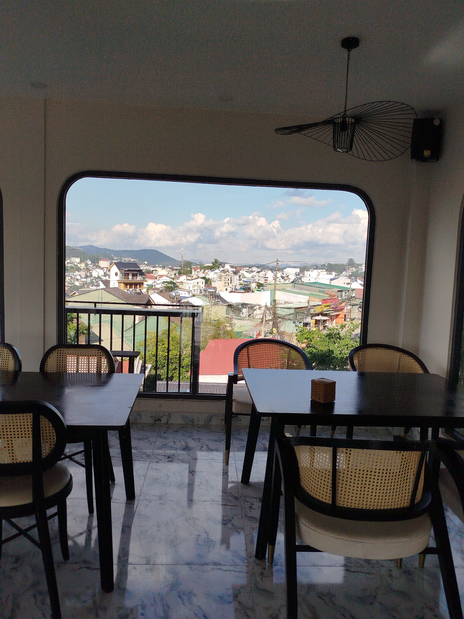 The image captures a cafe interior in Da Lat, Vietnam. Seating includes four tables with black frames and wicker chairs positioned near a large window overlooking the city. The view shows a cityscape with varied rooftops, trees, and distant hills under a blue sky with scattered clouds. A black, modern ceiling fan hangs above. The floor has a marble-like pattern. No people are visible.