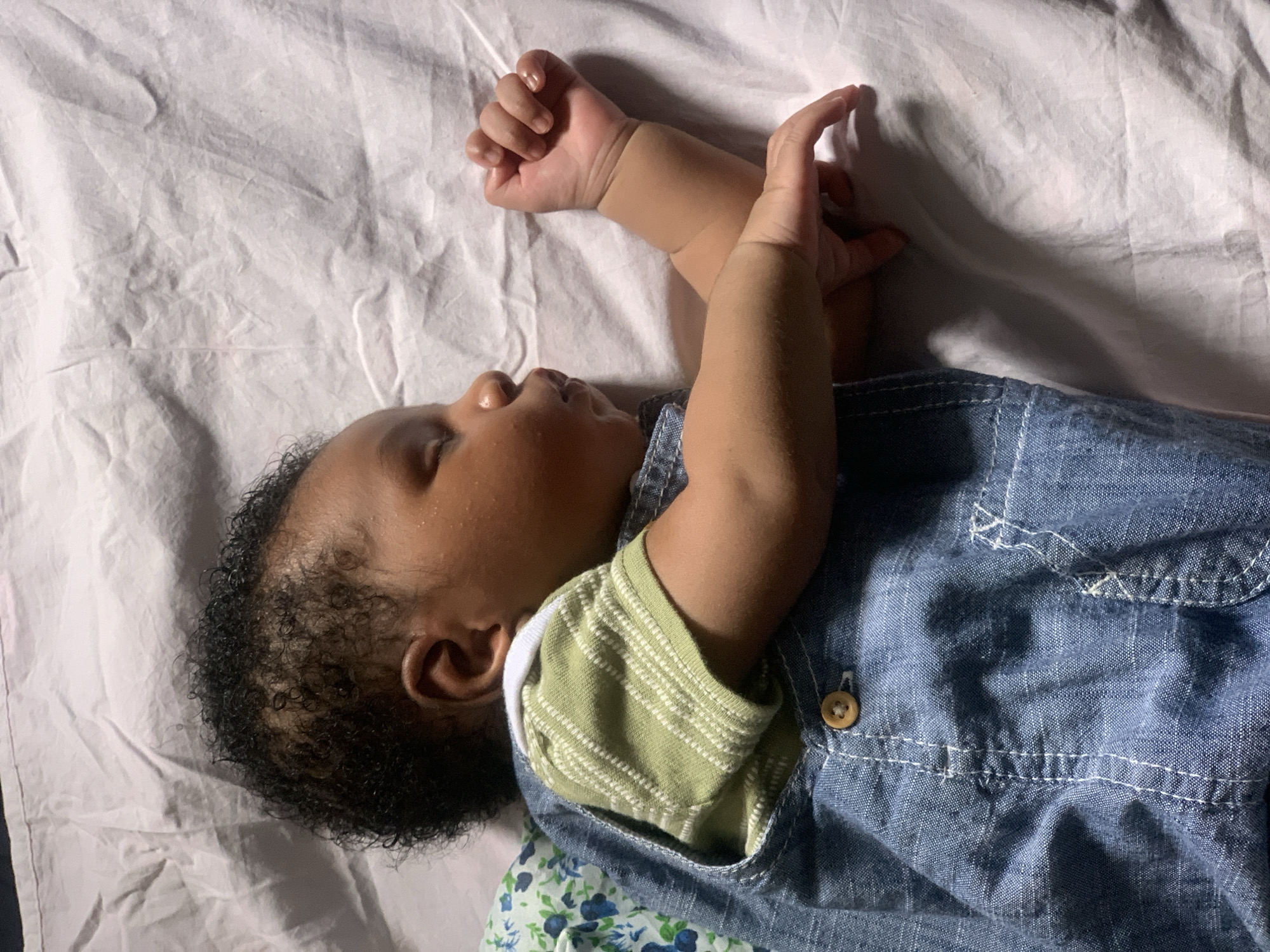 A baby is lying on a white sheet. The baby is wearing a green striped shirt under a denim overall. The baby's arms are raised with its hands near its face. The background is a white sheet. There is no evidence to support that this image was taken in Ibasa, Nigeria.