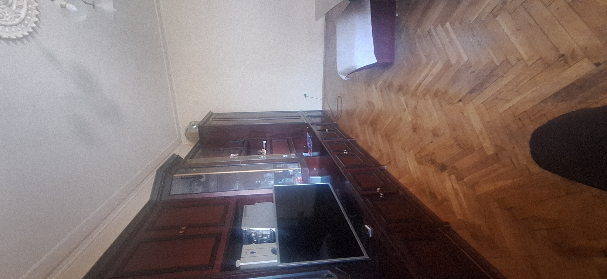 A high-angle view shows the interior of a room in Stara Zagora, Bulgaria. A large, dark-wood, built-in entertainment center is the dominant feature, including a television, glass shelves, and closed cabinets. A partially visible bed is in the upper-right corner. The flooring is parquet wood. A portion of a person's leg is visible in the lower-right corner. The ceiling has a decorative light fixture.