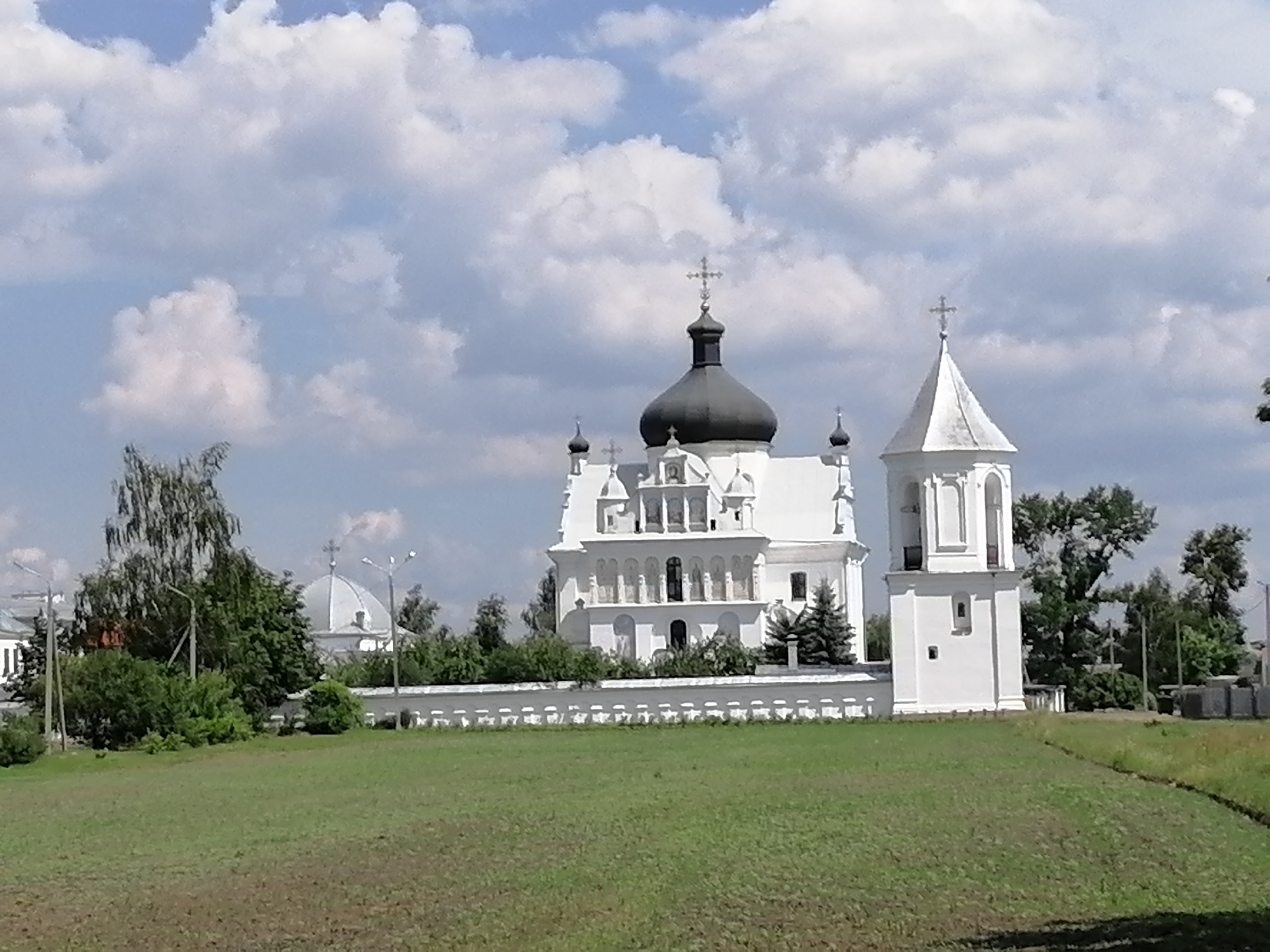 The media file shows a landscape view of a white, multi-domed church with a bell tower, set against a backdrop of a partly cloudy blue sky. The church has a large, dark dome topped with a cross, and several smaller domes, also adorned with crosses. The bell tower is white and rectangular with arched openings.
In front of the church, a low white wall runs along a green lawn. Beyond the wall, there are trees and some other buildings, including a dome-shaped structure to the left. A field of green grass occupies the foreground, with some darker patches suggesting plowed soil. The scene appears to be set during the daytime, with bright sunlight illuminating the church and casting shadows. The clouds indicate fair weather. The overall atmosphere is peaceful and serene. No people are visible in the image. There is no visible text in the media file.