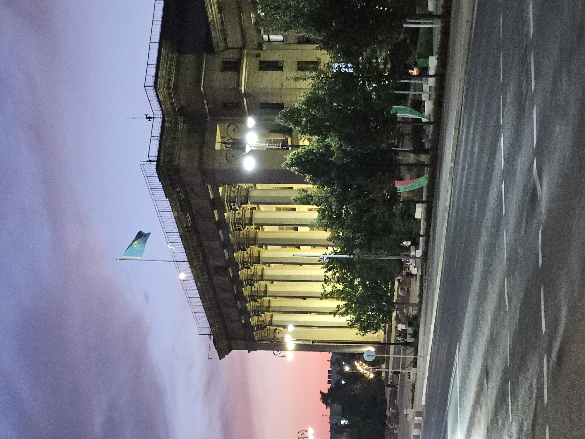 The image depicts a building with grand columns on its facade, likely a government or institutional building. The sky above is a gradient of purple and pink, indicative of twilight or dawn. A flag, appearing to be the flag of Kazakhstan, is visible on the rooftop, positioned to the left of center. In front of the building, there are several trees lining a walkway. The street in the foreground is dark and appears wet, reflecting some of the ambient light. Scattered individuals are present on the walkway and across the street, suggesting a public space. Decorative flags, green and red, are planted in front of the trees. The lighting suggests it is either early morning or late evening. The architecture of the building, particularly the columns and cornices, points to a classical or neoclassical style. This scene is consistent with the architecture and national symbols found in Almaty, Kazakhstan, specifically a prominent civic building.
