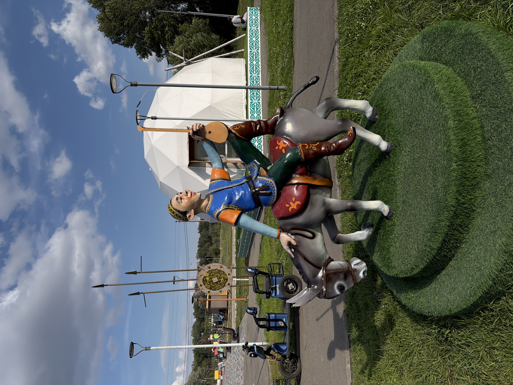 This is a full shot of a colorful statue of a man riding a donkey, set outdoors in a park environment. The statue is located in the city of Semey, Kazakhstan, possibly at a cultural or historical exhibit. The man, depicted in traditional attire, plays a stringed instrument. He wears a blue and orange vest over a light blue shirt, green leggings, and brown boots with ornate designs. The donkey is grey with a red saddle adorned with yellow and red symbols. The statue is positioned on a tiered green platform that resembles grass.
In the background, a large white geodesic dome structure with visible supports stands prominently. Several lampposts are visible throughout the park. To the left, a blue and black electric scooter and two blue wheeled carts are parked on a paved pathway. Further back, more park amenities like benches and pathways are visible, along with trees and a cloudy blue sky. In the distance, three upright spears with pointed tips are planted in the ground. A decorative circular emblem with green and yellow patterns is mounted on a stand behind the spears. The overall scene suggests a public outdoor space designed for leisure and cultural appreciation.