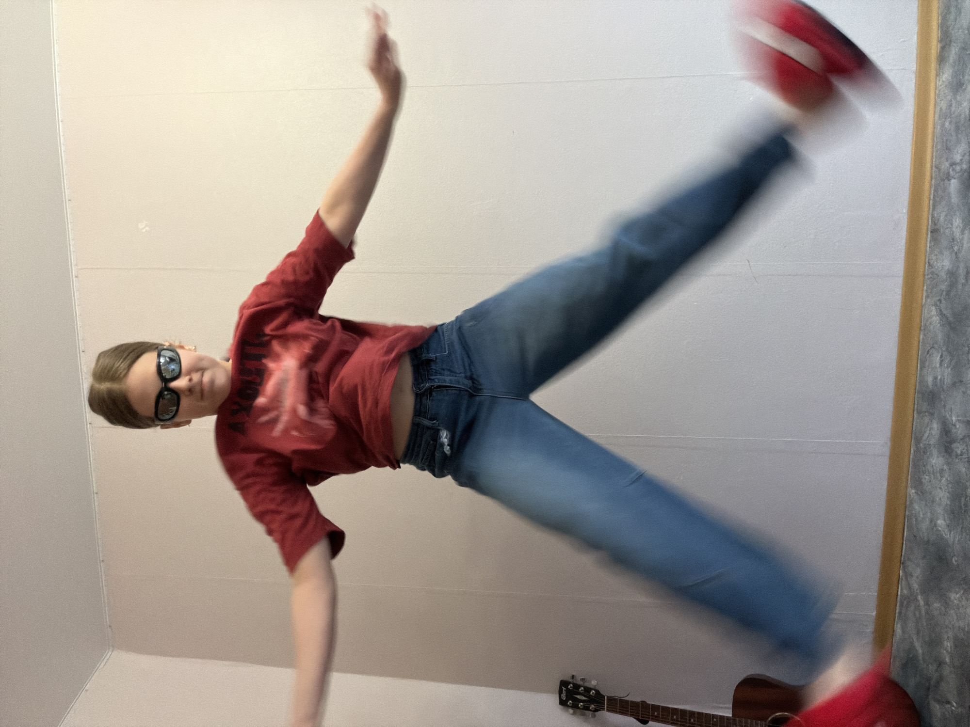 An image, captured at an angle, depicts a young person mid-action against a plain, light-colored wall. The individual, likely female, has light brown hair pulled back and wears black sunglasses, a red short-sleeved t-shirt with a patterned design, and light blue jeans. They are captured in a dynamic, acrobatic pose, with their left arm raised high and their right leg extended upwards and to the right. A red shoe on their right foot, along with their limbs, shows significant motion blur, conveying the speed of their movement.
The background consists of simple, off-white or light beige walls with subtle horizontal seams. In the lower right corner, a brown acoustic guitar is visible on the floor, partially obscured by the person's leg. The headstock of the guitar clearly displays the brand name "Cort." A darker, textured vertical surface runs along the far right edge of the frame. The indoor setting, likely a room or practice space in Perm, Russia, is illuminated by even, artificial lighting.