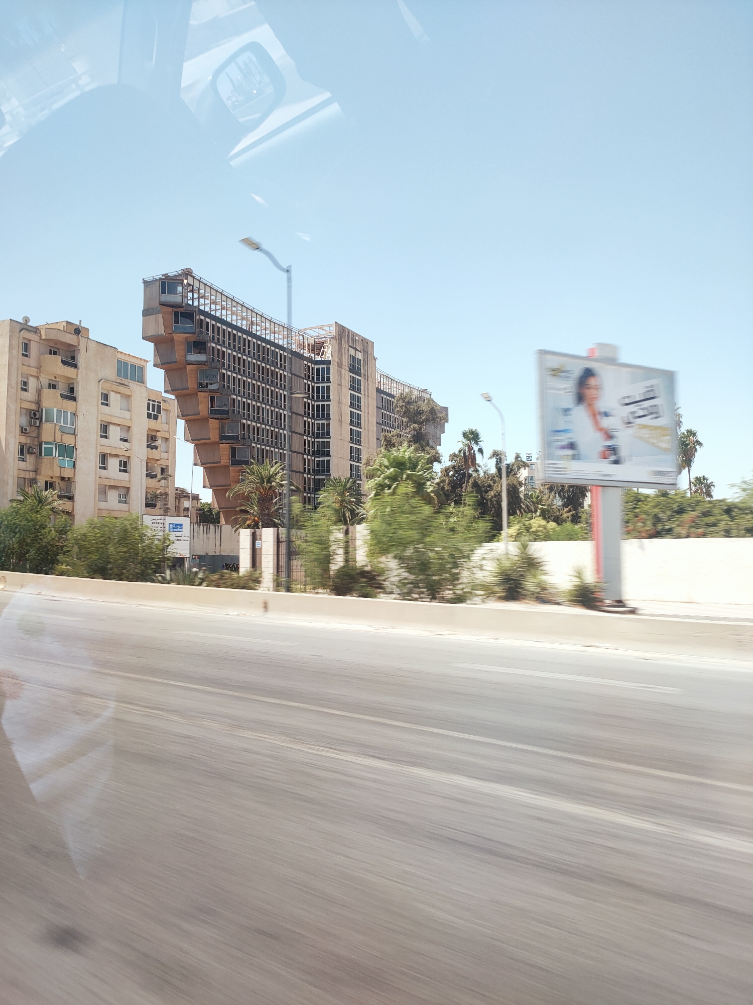 This is a street-level, medium-angle shot taken from inside a vehicle, showing an urban landscape in Tunis, Tunisia. The foreground is dominated by a blurred asphalt road, indicating movement. In the mid-ground, a distinctive Brutalist-style building with projecting concrete elements and numerous windows stands prominently. To its left, a lighter-colored apartment building with balconies and air conditioning units is visible. Palm trees and other greenery are scattered around the buildings. A billboard advertising a product or service with a woman's image is positioned on the right side of the frame, also showing motion blur. The sky above is clear and pale blue. The reflection of the car's interior, including a rearview mirror, is visible on the left side of the image, overlaying the scene.