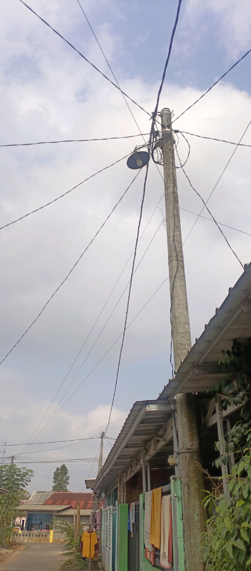 In Kotabumi, Indonesia, a street scene unfolds beneath a cloudy sky, dominated by a concrete utility pole supporting a street lamp and a complex network of electrical wires. The pole stands tall, its surface weathered and marked, with wires intricately woven around it. The street lamp, a dark, saucer-shaped fixture, is attached to the pole, its bulb visible. Adjacent to the pole, the roofline of a building stretches into the frame, its corrugated metal surface glinting. Further down the street, houses with red-tiled roofs are visible, interspersed with smaller structures and foliage. In the foreground, on the left side of the image, a yellow shirt hangs from a clothes rack. The scene suggests a residential area with overhead utility infrastructure.