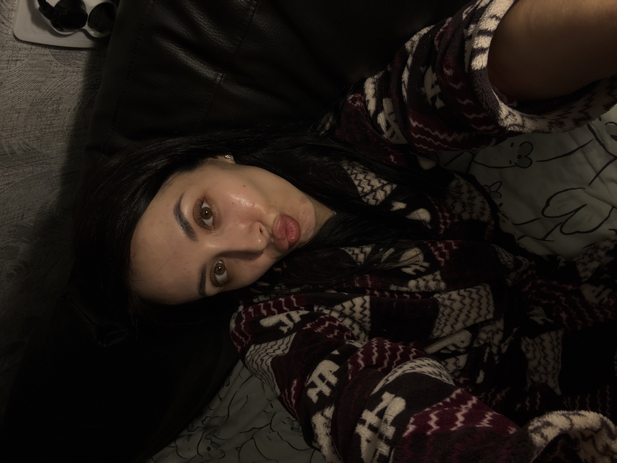 A selfie photograph presents a woman lying down in what appears to be an indoor setting, potentially a residence. She has dark hair and brown eyes, and is wearing a patterned robe or sweater. She is making a pouting facial expression, a common posture in selfies. The background suggests she is resting on a dark-colored sofa or leather surface, with a patterned blanket or bedding underneath. There is an indistinct object in the upper left corner of the frame, possibly headphones. The photograph's perspective is from above, as if she is holding the camera and pointing it down at herself. The setting is not explicitly identifiable as Votkinsk, Russia, and no other definitive information about the location is present.