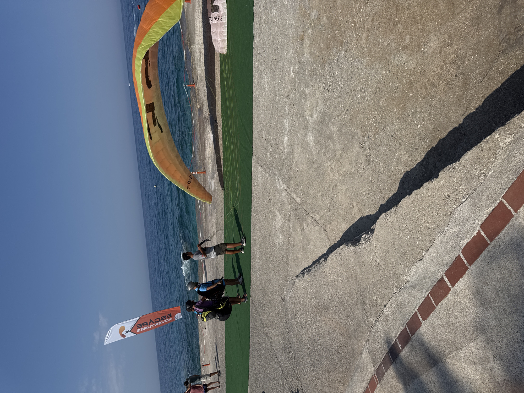 In Fethiye, Turkey, on a sunny day, several individuals are gathered on a concrete platform near the sea, with paragliders and equipment present. A tall, orange and white flag with the text "ESCAPE ADVENTURES" is planted in the background. A large paraglider wing, colored orange and green, is spread out over a green mat on the platform, partially inflated. Three people are standing on the green mat, one wearing a helmet and backpack, another in a blue shirt and shorts, and a third in a gray t-shirt and shorts, holding paragliding lines. In the distance, the blue sea meets a clear sky. The concrete platform has a visible expansion joint and is bordered by a red-brick pathway in the foreground. The overall scene depicts preparations or a break during paragliding activities by the coast.