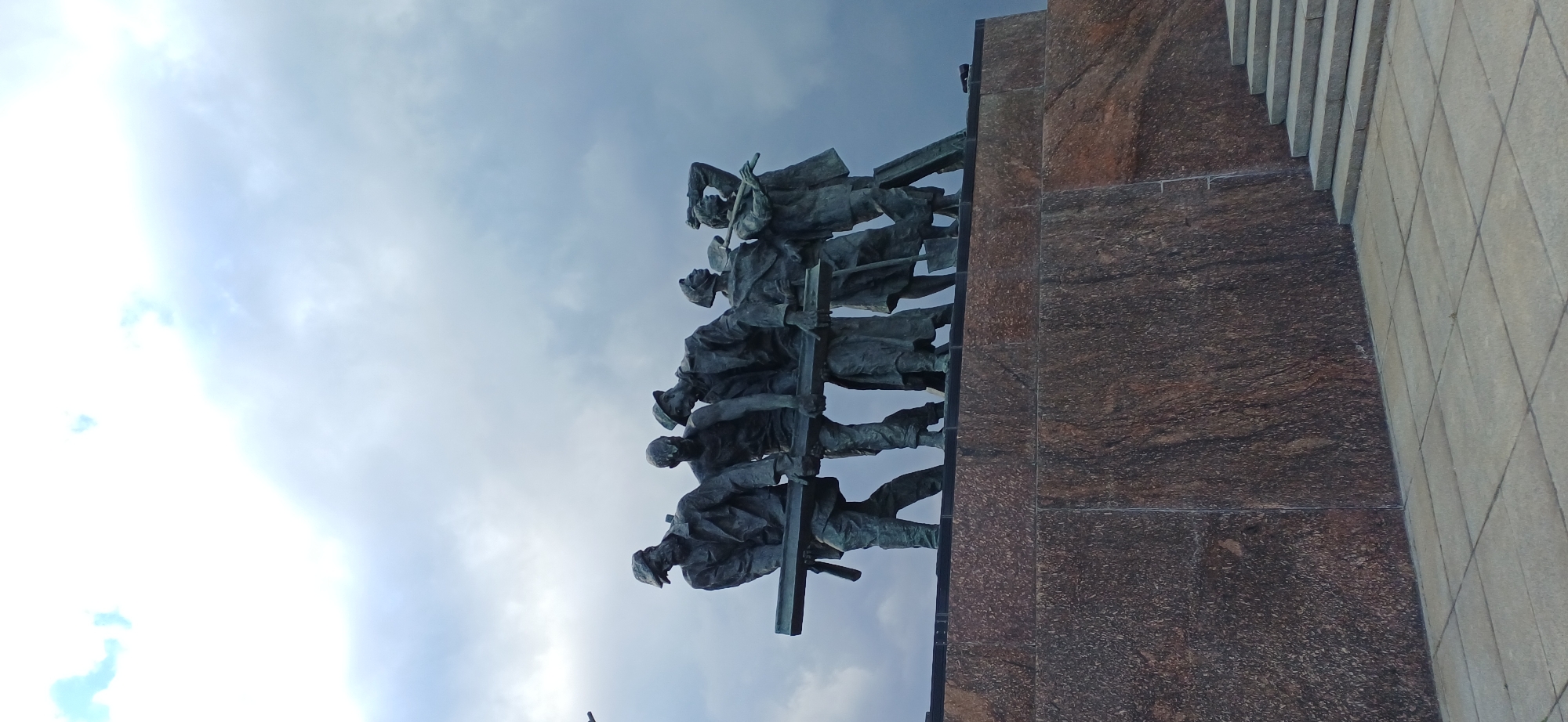 The image shows a bronze sculpture of six workers carrying a large beam. The sculpture is positioned on top of a stone pedestal. The workers are depicted in a collective effort, with their bodies straining to support the weight. They are dressed in clothing that suggests a historical or industrial setting, possibly from the Soviet era. The sky behind the sculpture is dramatic and overcast, with dark clouds suggesting an impending storm. The lower part of the image shows a stone staircase leading up to the left, and a paved area in the foreground. The sculpture is likely located in Saint Petersburg, Russia, and given its theme, it could be part of a memorial or monument related to labor, industry, or historical events in the city.