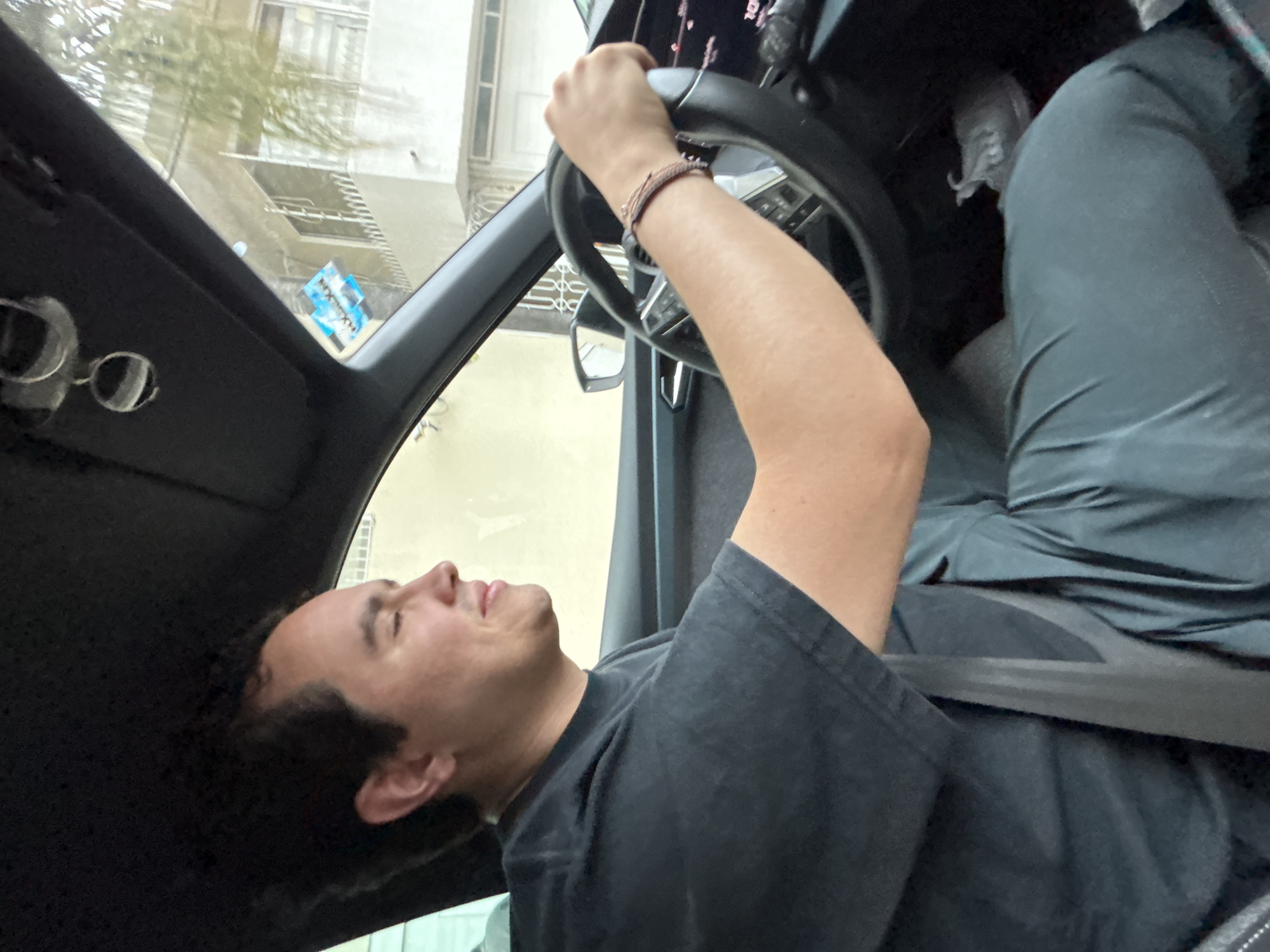 A male is sitting in the driver's seat of a car, holding the steering wheel. He is wearing a dark gray t-shirt and a multi-strand bracelet on his wrist. He has dark hair and his eyes are closed. A seatbelt is across his chest. Outside the car window, a building is visible. The image appears to be taken from inside the car. The location is in Puebla, Mexico.