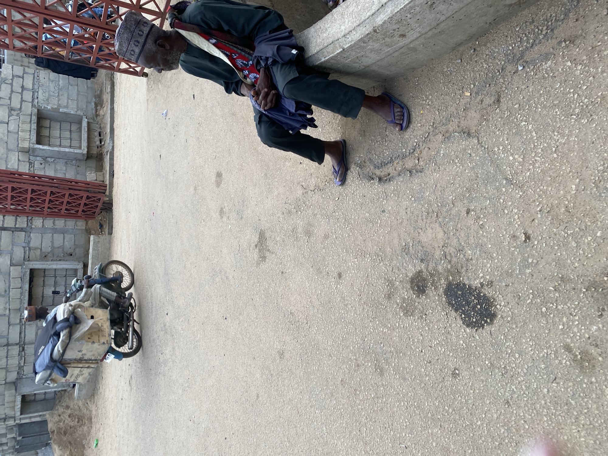 In Jos, Nigeria, an elderly man with dark skin and gray hair sits on a concrete barrier, facing right. He wears a dark green robe, dark pants, a patterned skullcap, and purple flip-flops. A colorful cloth is draped over his shoulders, and he holds a dark blue cloth in his lap with clasped hands. To the left, a person on a motorcycle drives past, carrying a large load of items secured in a box and plastic wrap. The motorcycle is blue and gray. The scene takes place on a paved road in front of a building under construction, with a red metal gate visible in the background. The environment appears to be an outdoor urban setting.