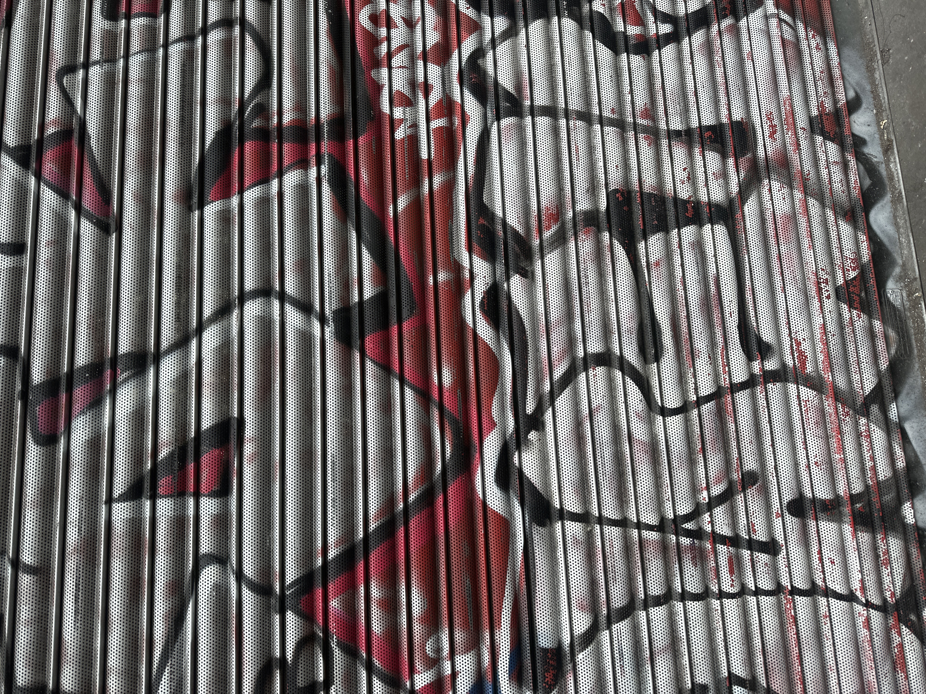 The image displays a perforated metal roller shutter, likely from a shopfront, situated in the City of London, United Kingdom. The shutter is heavily covered in graffiti, primarily featuring bold abstract designs in red and black spray paint. The red appears to be an underlying fill, with prominent black outlines and shapes layered on top. The perforations of the metal shutter are clearly visible through the paint application, creating a textured, dotted appearance. The horizontal slats of the shutter are distinct. At the bottom, the shutter meets a grey, possibly concrete, ground surface that shows minor dirt and debris. No individuals, animals, or additional architectural features are visible beyond the immediate context of the shutter and ground.