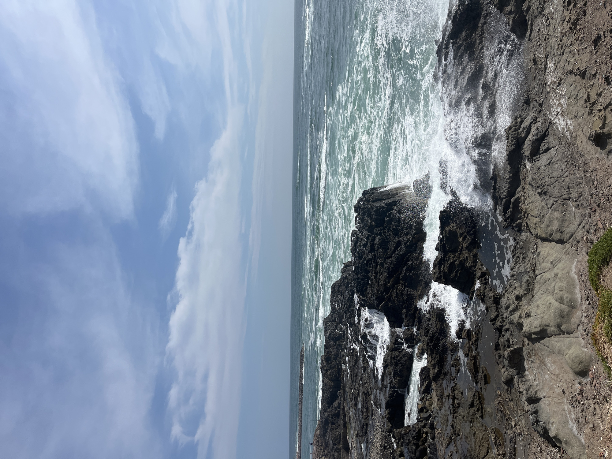 The image depicts a rocky coastline in Arica, Chile. The view is of the Pacific Ocean crashing against dark, jagged rocks, creating white foam and spray. The ocean stretches to the horizon. To the left, the sky is partly cloudy with patches of blue. The photograph is taken from an elevated position, providing a broad view of the coast. A jetty is visible in the distance, slightly left of center. No people are visible in the frame.