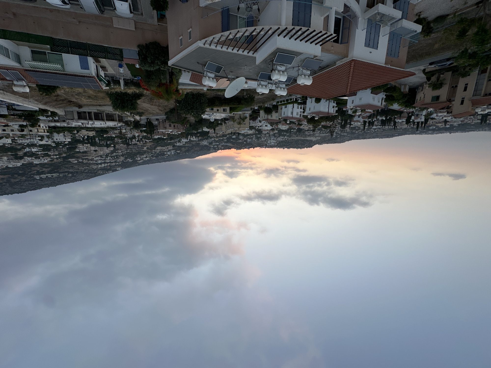 This is a high-angle, wide shot of Peyia, Cyprus at dusk. The sky is filled with a mixture of pale blue and scattered clouds, with hints of pink and orange from the setting sun near the horizon. The landscape is dominated by a hillside densely populated with white and light-colored buildings, interspersed with trees and sparse vegetation. In the foreground, a multi-story building with a terracotta tiled roof is visible, featuring blue shutters on its balconies. On the roof of this building, there are several solar water heaters and solar panels, along with a satellite dish. Further into the scene, more residential buildings are spread across the hillside, and a road can be seen winding through the town. The overall impression is a quiet residential area under a dramatic, colorful sky.