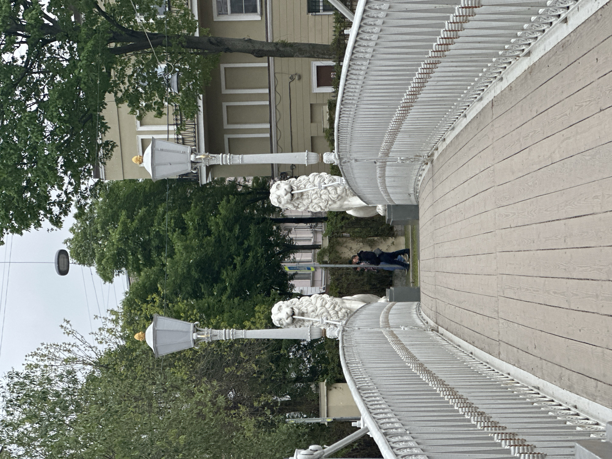 The image shows a segment of the Lion Bridge (Lviny Most) in Saint Petersburg, Russia. The bridge is white with a wooden walkway. Two white lion sculptures are visible, each holding a lamppost. The lampposts have gold-colored finials. The bridge railings are white and ornate. In the background, there are green trees and a light-colored building with vertical windows. There are also two individuals walking in the background. Overhead wires are present. The lighting suggests an overcast day.