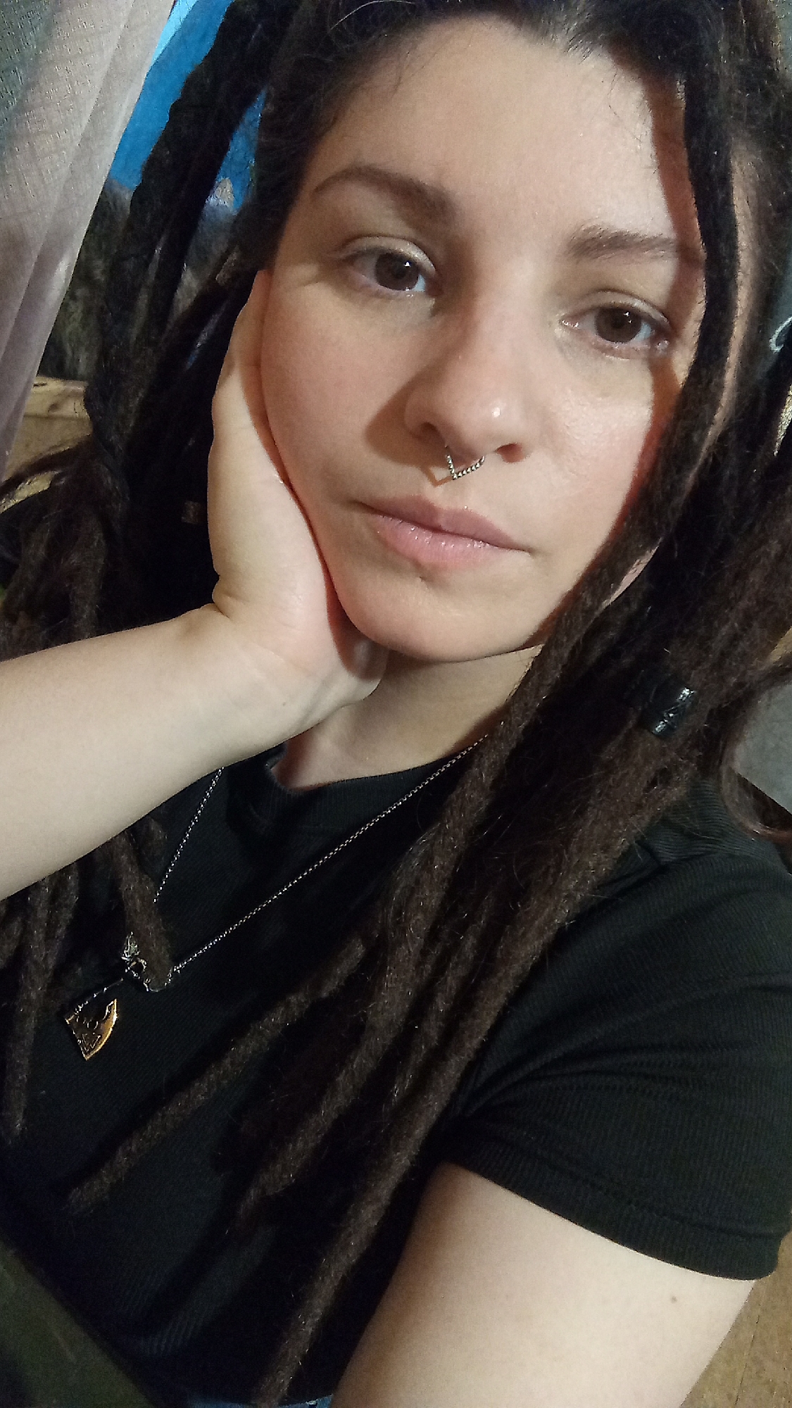A medium-shot photograph features a person with light skin, dark brown dreadlocks, and a nose ring, resting their left cheek on their left hand. The person is wearing a black ribbed t-shirt and a silver necklace with a pendant. The background is blurred, showing hints of light blue, white, and green colors. The location is unknown, as the image does not provide any clues indicating Юдино, Russia.