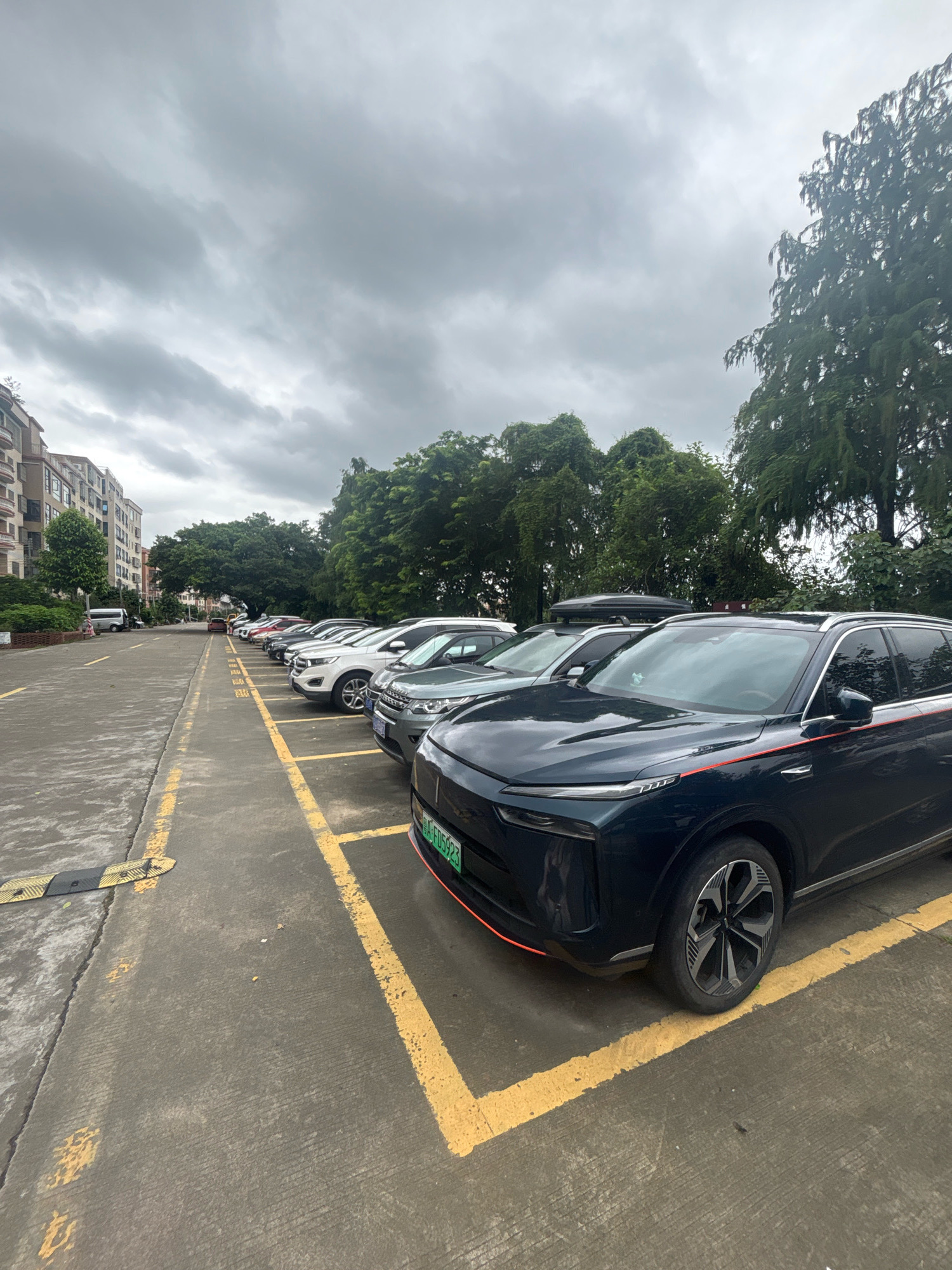 A line of parked cars is visible in a parking lot in Nanhai District, China. The cars are positioned in marked parking spaces with yellow lines. The foreground features a dark blue SUV with red trim, its front wheel clearly visible. Further back, a row of other vehicles, including white and silver SUVs, are parked. To the left of the parking lot, a multi-story residential building is situated, with lush green trees bordering the scene. The sky above is overcast with gray clouds. No people are clearly visible interacting with the vehicles.