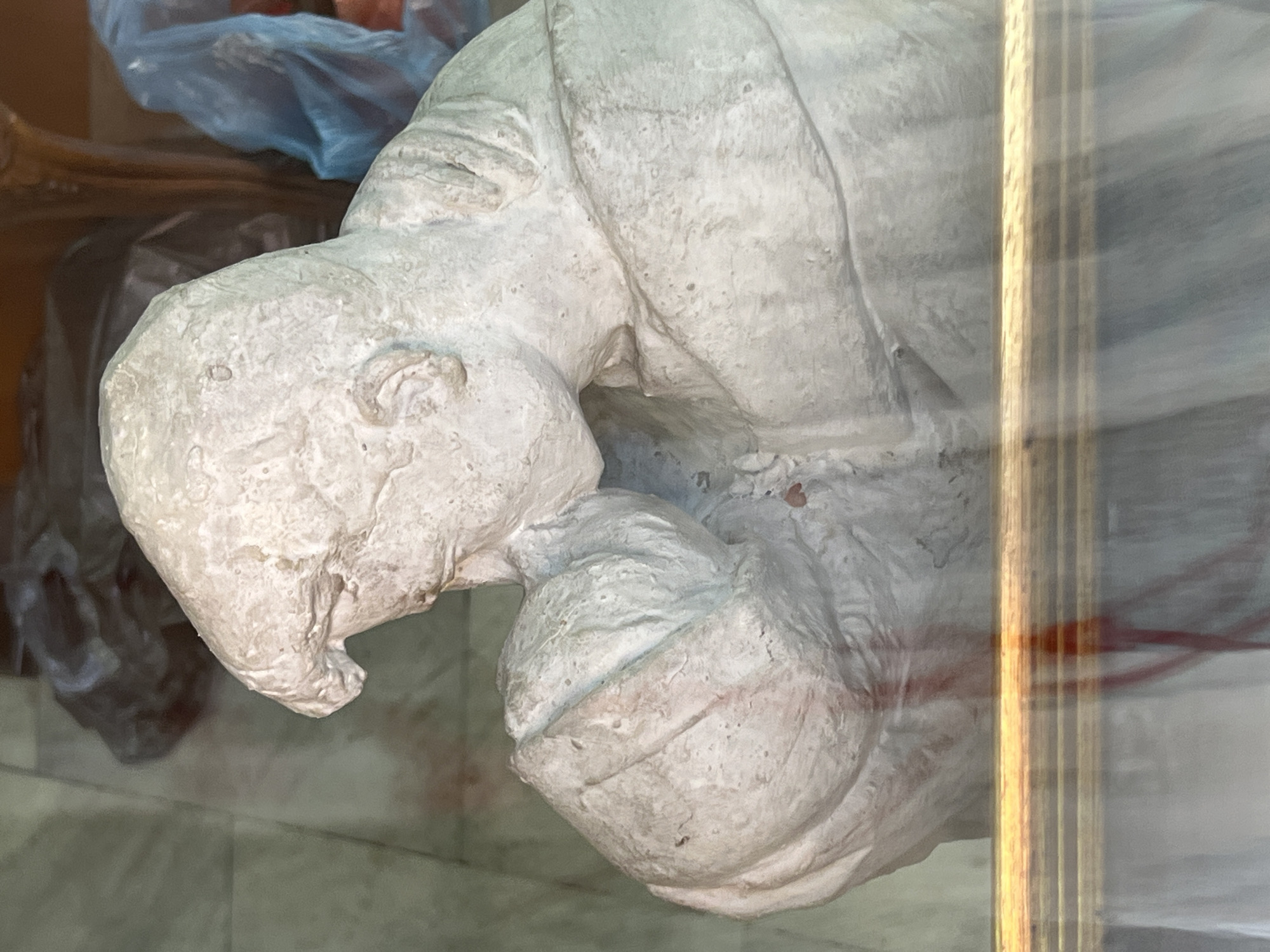 The image shows a close-up view of a plaster sculpture of a person, possibly a bust, with the head tilted down and the hands brought up towards the face, creating a posture of contemplation or distress. The sculpture is light-colored, with visible texture and some reddish smudges or stains on its surface, particularly on the chest area and the right shoulder.
The sculpture is positioned behind a glass surface, indicated by reflections and a horizontal golden bar across the lower part of the image, which appears to be part of the display or enclosure. In the background, through the glass and behind the sculpture, various objects are visible, including what appears to be crumpled plastic bags (one dark, one blue) and a hint of a wooden structure. The flooring in the background seems to be tiled.
The provided context specifies the location as Sofia, Bulgaria. Without further information about the specific exhibit or venue, it is impossible to pinpoint the exact location within the city. However, such sculptures are commonly found in museums, art galleries, or private collections. The presence of what appears to be storage materials in the background could suggest a workshop, storage area, or a less curated exhibition space.
