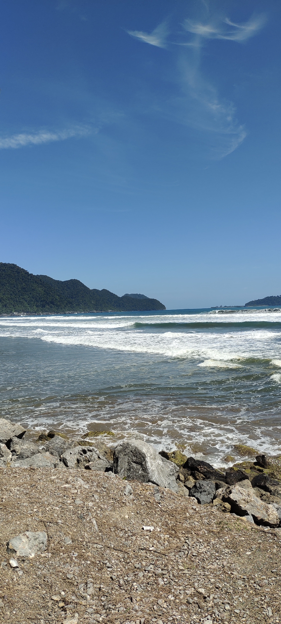 A vertical, eye-level shot captures a serene beach scene with lush green mountains in the background under a clear blue sky. The foreground is a rough, gravelly embankment with scattered rocks. Beyond the embankment, the ocean stretches out, its surface dotted with white waves. The water is a mix of blues and greens, with foamy whitecaps. In the distance, two prominent green, forested islands form a natural boundary, with a hint of man-made structures visible on the far right island. The sky is a vibrant blue, with a few wispy white clouds adding texture. The overall impression is one of natural beauty and tranquility.