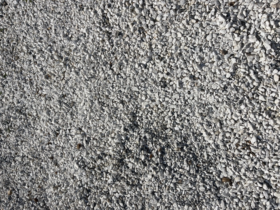 The image shows a ground covered in white gravel. The gravel is densely packed, with minimal space between the stones. The gravel is a mix of small and medium-sized white stones, with some darker grey stones interspersed throughout. The gravel is illuminated by sunlight, which highlights the texture of the stones and creates a sense of depth. The image was taken in Limenaria, Greece, at latitude 40.626, longitude 24.57. The image does not contain any people or objects.