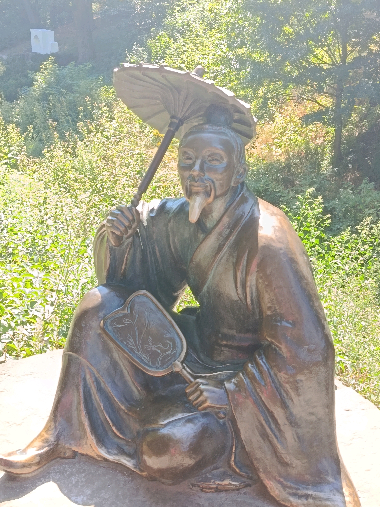 This is a bronze statue of an elderly Asian man sitting cross-legged outdoors, holding a traditional fan in his left hand and an umbrella in his right. He has a long, white beard and is wearing a flowing robe. The statue is positioned on a stone base amidst lush green foliage, with trees visible in the background under a bright, sunlit sky. A white, abstract sculpture is faintly visible in the upper left background. This statue is located in the Dendropark Oleksandriya in Bila Tserkva, Ukraine.