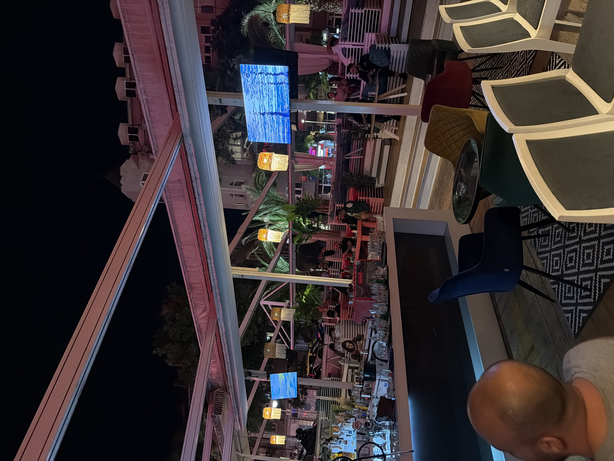 This is a detailed, matter-of-fact description of an evening scene at an outdoor bar or restaurant in Plovdiv, Bulgaria.
The image captures a vibrant night setting illuminated by a combination of artificial lights. A large, open-air structure, likely an awning or retractable roof system, dominates the upper part of the frame, with visible white and reddish-orange beams against the dark night sky. In the background, urban buildings are visible, including one with a distinct pointed roof and multiple dormer-like windows, characteristic of European architecture. Lush green foliage, including palm trees, is integrated into the establishment's decor.
In the foreground to the left, a well-stocked bar counter is visible, displaying various liquor bottles, including gin, along with glassware and an ice bucket. One individual, seen from behind with a bald head and wearing a grey t-shirt, is seated at the bar.
The central and right portions of the image feature multiple tiered seating areas with a variety of chairs and tables. Seating options include blue velvet chairs, yellow quilted chairs, red velvet chairs, dark green chairs, and light-colored chairs with grey cushions. Small, dark circular tables, some equipped with ashtrays, are interspersed among the seating. Patterned rugs are placed beneath some of these areas.
Several patrons, estimated to be between 10 and 15 individuals, are scattered throughout these seating zones, primarily in small groups or pairs. They appear to be engaged in conversation or looking at mobile devices.
Two large electronic screens are prominent features of the decor, displaying ambient blue patterns; one shows a cloudy sky, and the other depicts dynamic water ripples. Decorative hanging lamps with woven, fringed shades emit a warm, inviting glow throughout the space. The overall environment suggests an active, relaxed evening social setting.