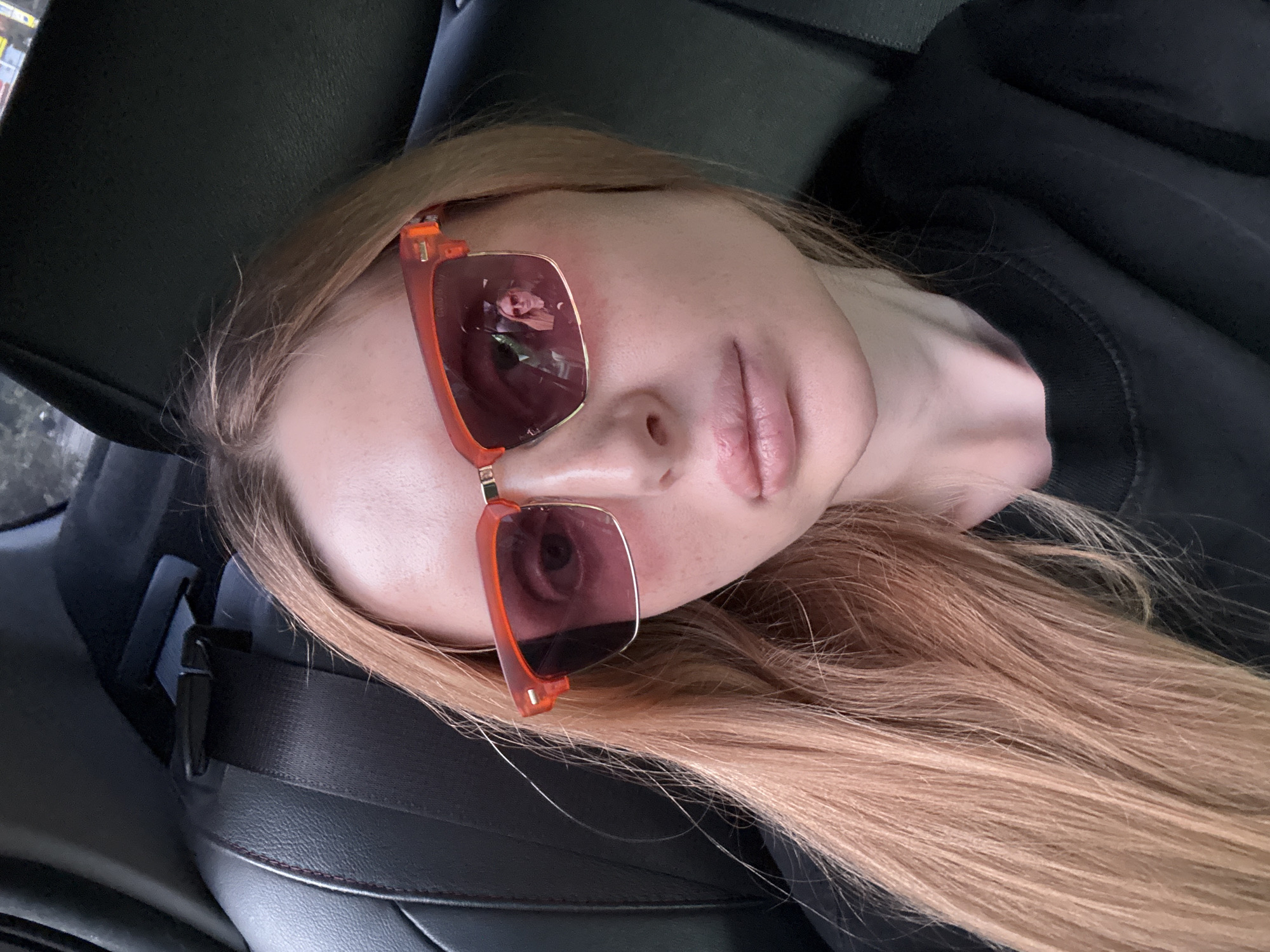 A close-up, high-angle, sideways shot shows a young woman wearing orange-framed sunglasses with pinkish lenses. Her face is angled upwards, with her mouth slightly parted. Her long, light brown hair is spread out across the frame. The reflection in the sunglasses shows her face and the interior of a vehicle. She is wearing a black top. The background consists of dark car interior elements, including a headrest and a seatbelt buckle. The scene appears to be indoors or inside a vehicle. The lighting suggests it might be daytime. No visible text is present.