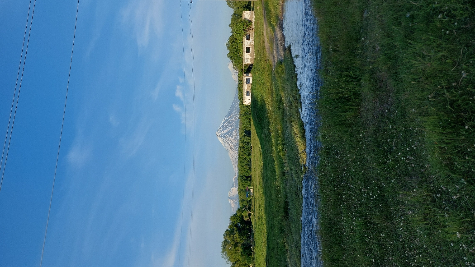 This vertical, medium-shot photograph depicts a natural landscape in Koryaki, Russia, under a clear blue sky with thin, wispy clouds. In the foreground, a shallow, winding stream flows across a grassy field dotted with small white flowers. Tall, lush green grass lines the banks of the stream, and the field itself is verdant. In the middle ground, a line of dense green trees forms a backdrop to a dilapidated, rectangular building with several open, dark window frames. The building appears to be constructed of concrete blocks and is in a state of disrepair. Behind the trees and the building, a majestic, snow-capped volcanic mountain range dominates the horizon. The largest volcano, in the center of the frame, is conical and prominently snow-covered, suggesting a cold climate or high altitude. Two smaller, also snow-covered mountains are visible to its left and right. Two thin, parallel power lines stretch horizontally across the top portion of the image. The scene suggests a rural or undeveloped area, likely in the early morning or late afternoon given the soft lighting. There are no people or animals visible in the image.
