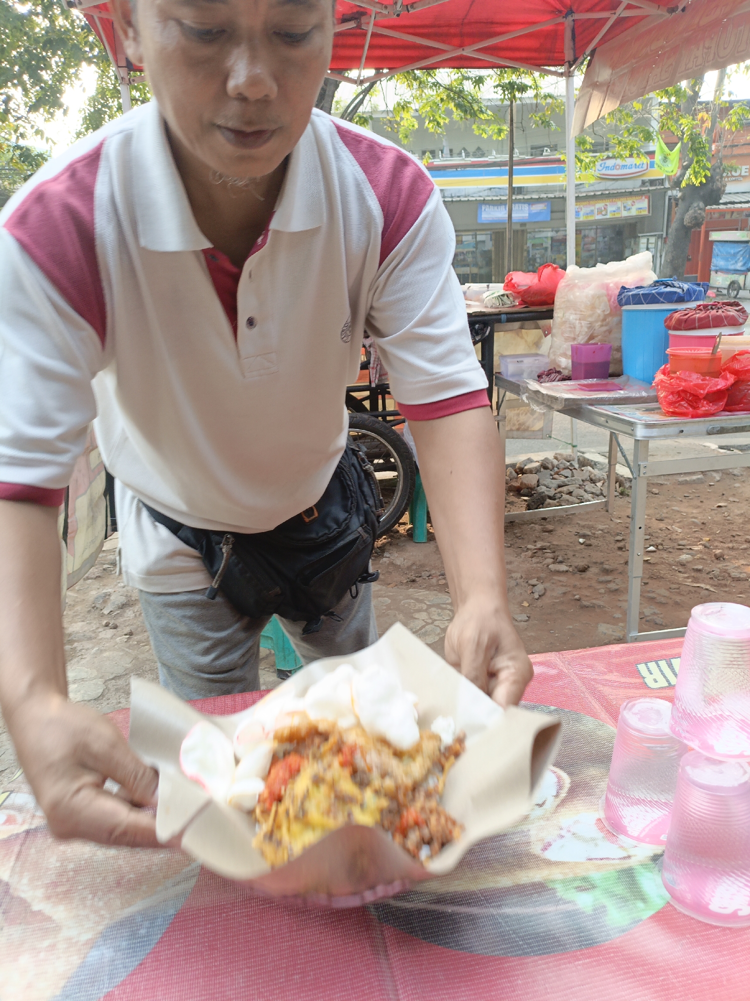 A person is standing behind a table at an outdoor food stall in East Jakarta, Indonesia. The person, wearing a white polo shirt with pink accents and grey sweatpants, is holding a serving of food wrapped in brown paper. The food appears to be a mix of colorful ingredients and white crackers. On the table are various items, including bags of snacks, plastic cups, and containers. In the background, there are buildings, trees, and a red canopy above the stall. The ground is unpaved and rocky. The overall scene depicts a vendor preparing food for a customer.
