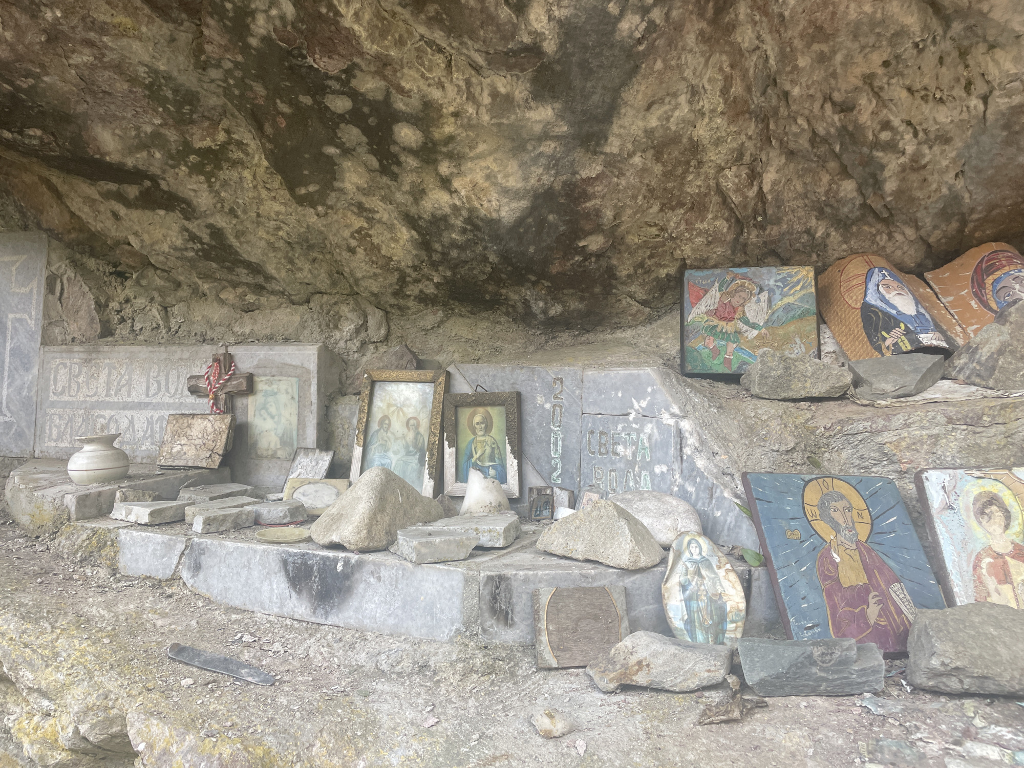 The image depicts a small, open-air religious shrine nestled in a cave-like rock formation. This is likely a roadside shrine in a rural or natural setting.
Several religious icons, paintings, and objects are arranged on a stone ledge, serving as an altar. These include images of saints and religious figures in various styles and formats, some appearing to be painted on stone or wood. There are framed pictures, stone tablets with inscriptions (possibly Cyrillic), a small vase, and a cross. Rocks and small stones are scattered around the altar, apparently used to prop up or secure the icons.
The setting is natural, with the rocky cave backdrop providing a rough and uneven background. The lighting is subdued, suggesting the shrine is located in a shaded area. The overall impression is one of a rustic, simple, and well-maintained place of worship or devotion.
There is no confirmation in the image that this scene is set in Sofia, Bulgaria.