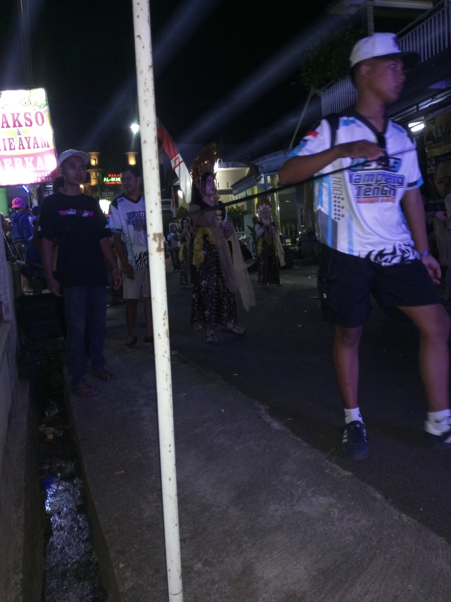 The image depicts a nighttime scene on a street in Langlang, Indonesia. A group of people are participating in what appears to be a parade or cultural event. In the foreground, a person wearing a white baseball cap and a white, blue, and black patterned t-shirt with black shorts is walking, facing to the right. To the left of this person, another individual in a black t-shirt and blue jeans is also walking. Further back, several other individuals are visible, some in traditional attire. One person in the background wears a distinctive headdress and a patterned dress. A red and white flag is also visible among the participants. The environment suggests a built-up area with buildings and streetlights. A drainage ditch runs along the side of the street in the foreground, containing some debris. The lighting indicates artificial illumination, likely from streetlights and possibly from the event itself.