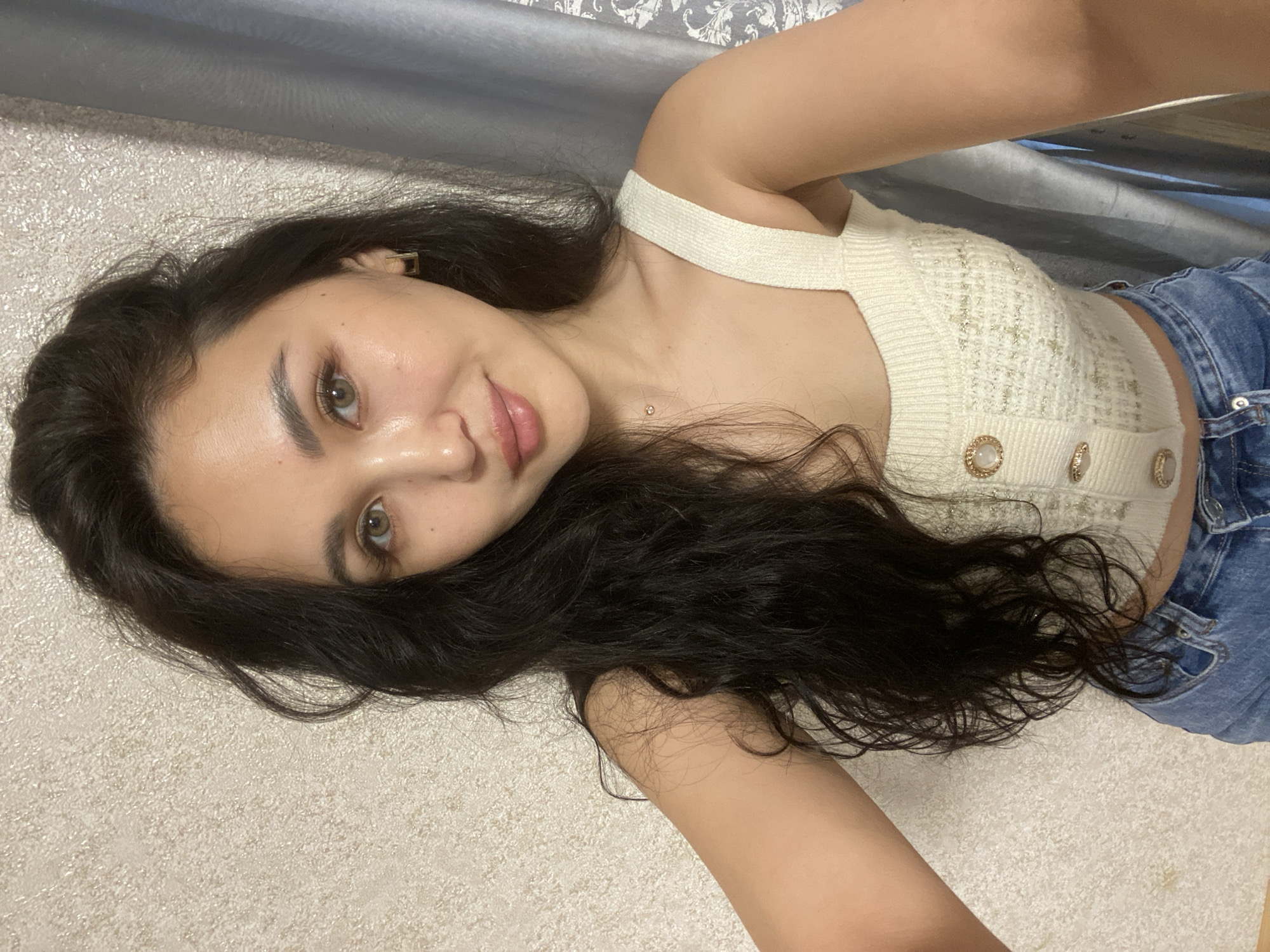 A woman with long, dark hair and olive skin is posing in a selfie-style shot. She is wearing a light-colored, textured tank top with decorative buttons, and blue jeans. The background consists of a light-colored, textured surface and a blurred grey area suggesting a wall or curtain. The location cannot be determined based on the image.