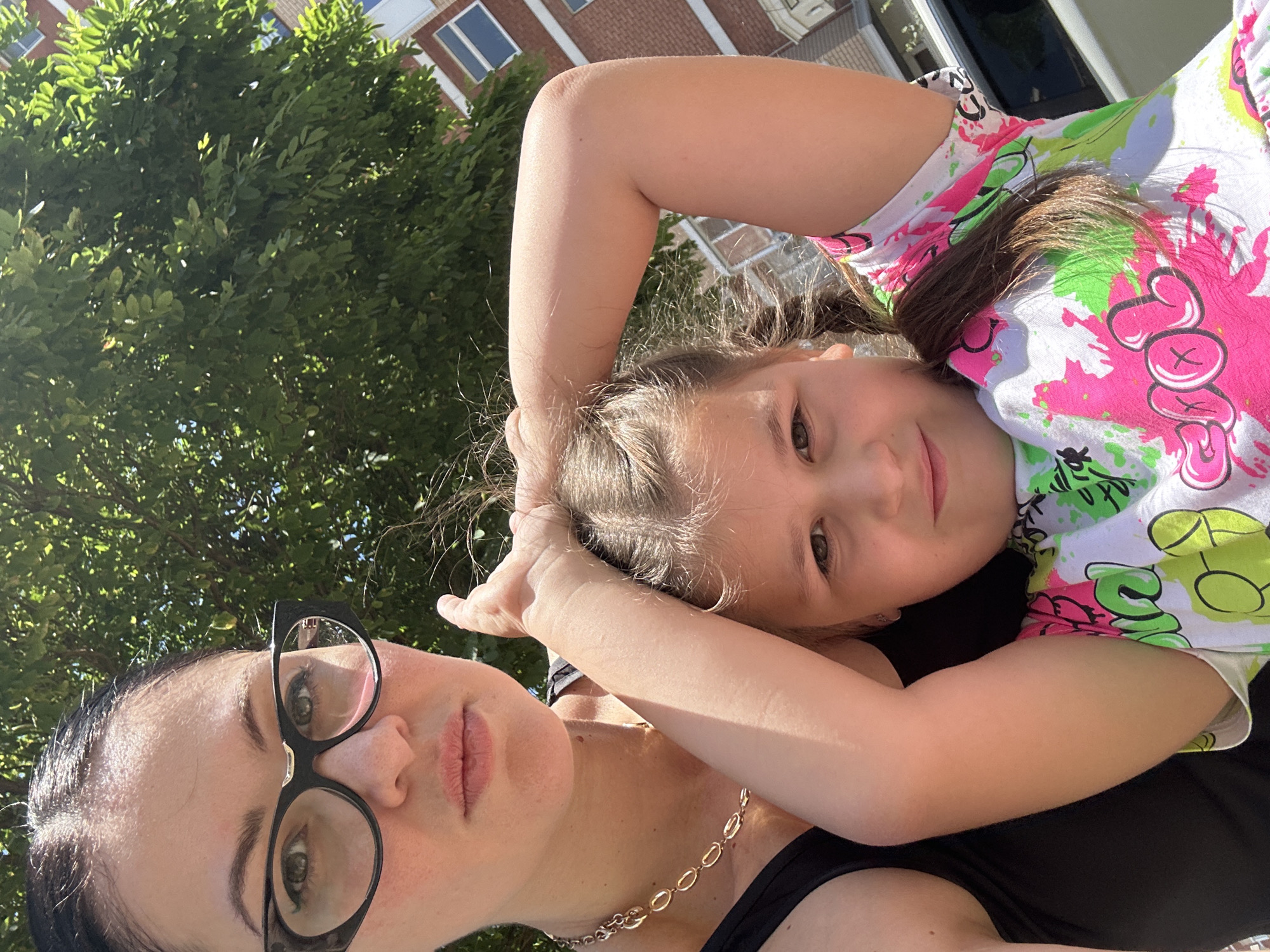 A selfie taken outdoors shows a woman and a young girl. The woman, wearing black glasses and a black tank top, is in the foreground on the left. She has dark hair and is looking at the camera. Next to her, slightly in front and to the right, is a young girl with braided brown hair. She is wearing a white t-shirt with colorful abstract patterns and is holding her arms up behind her head. Sunlight illuminates both subjects. In the background, there is a large green tree with dense foliage, and a brick building with windows can be seen. The scene suggests a sunny day in a residential or urban area.