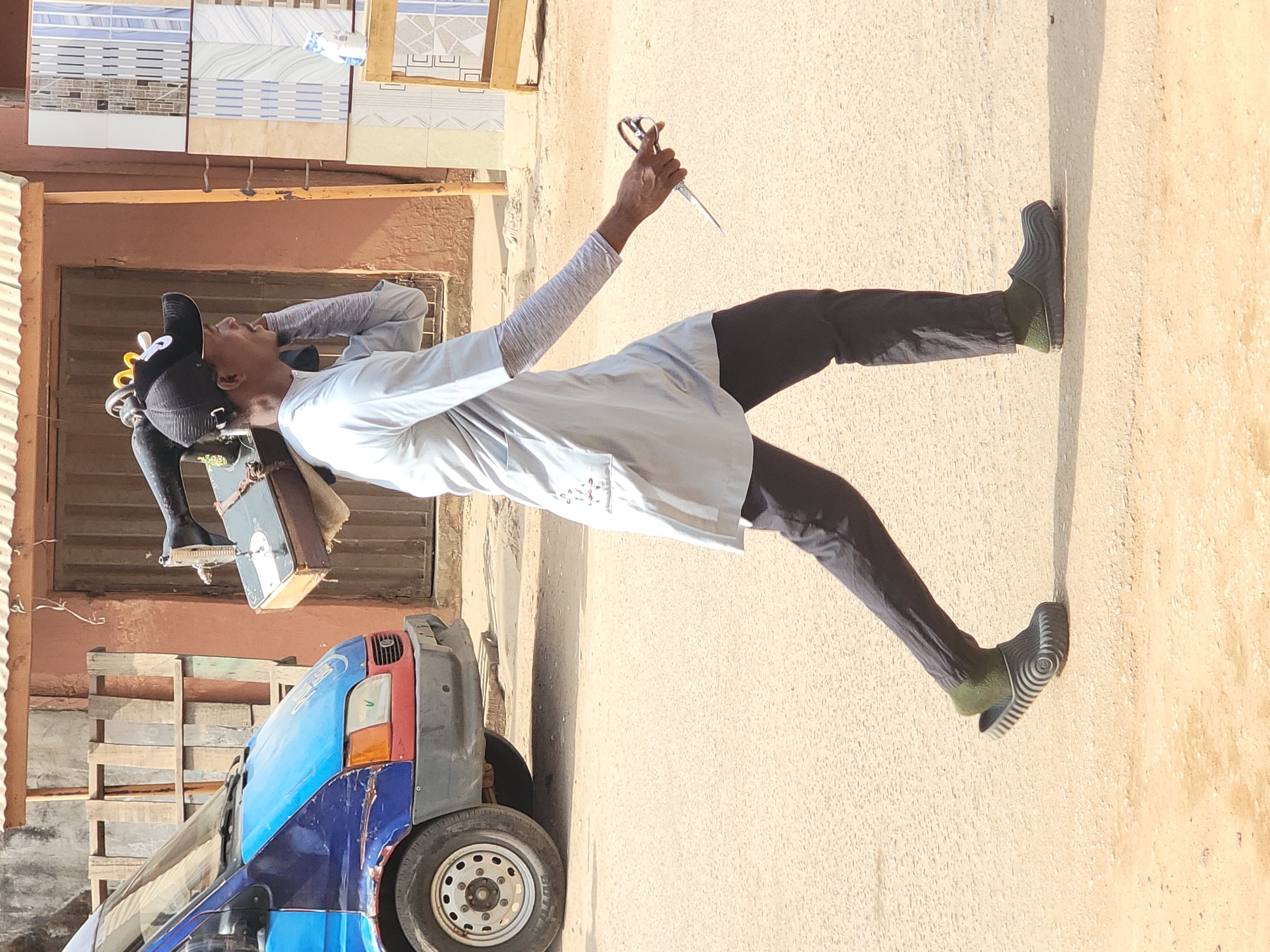 The image, taken in Maiburiji, Nigeria, captures a man standing outdoors on what appears to be an unpaved street or market area during bright daylight. Strong sunlight casts distinct shadows on the ground and walls.
The main subject is a man dressed in a light-colored, long-sleeved shirt and dark trousers, paired with dark grey slip-on shoes featuring green soles. He wears a black cap, under which several hair curlers or similar styling tools are visible on his head. In his raised right hand, he holds a pair of large, sharp scissors or shears. His left arm is bent, with his hand near his head, possibly adjusting his cap, touching his hair, or shielding his eyes from the sun.
A vintage-style portable sewing machine, mounted on a stand or box, is positioned to his left, indicating he might be a tailor or involved in garment work. In the lower left background, the front section of a blue and red van or small bus is partially visible.
The background includes a display of various building materials, specifically several samples of wall tiles with different patterns (striped, geometric, and textured designs) mounted on a structure, suggesting a nearby shop or stall selling such items. Wooden pallets or crates are also present in the background. The ground is a light, sandy color, contrasting with a light-colored wall.
No prominent text is visible on any of the subjects or objects in the scene. The image depicts a slice of daily life and outdoor commerce.