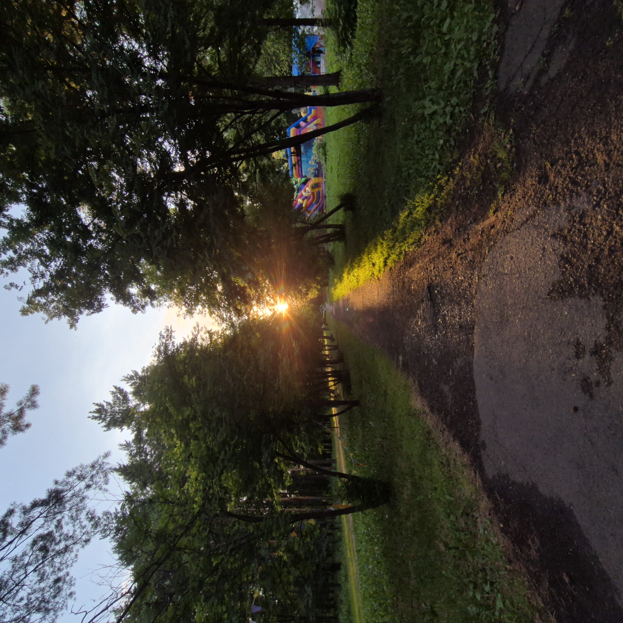 This image depicts a sun-drenched path bordered by trees and lush green grass in Kemerovo, Russia. The sun is low in the sky, casting long shadows and creating a warm, golden glow across the scene, suggesting it is either early morning or late afternoon. The pathway appears to be asphalt or a similar paved surface, and it curves gently into the distance, disappearing among the trees.
On the right side of the path, the ground is a mix of dark soil and gravel, with some dry leaves scattered about. The grass on the left side of the path is vibrant and green, with patches of sunlight highlighting its texture. In the background, through a gap in the trees, colorful inflatable structures, possibly part of a playground or entertainment area, are visible. These structures are a mix of bright blues, yellows, reds, and purples, adding a pop of color to the otherwise natural setting.
Several trees line both sides of the path, their leaves appearing full and healthy. The sun's rays are bursting through the branches, creating a lens flare effect and illuminating a section of the path with bright light. Small, colorful flags or pennants are strung between some of the trees along the path, adding a festive touch to the environment.
The overall mood of the image is serene and peaceful, with the warm sunlight evoking a sense of tranquility. The presence of the inflatable structures hints at a public recreational space. There is no visible text in the image.