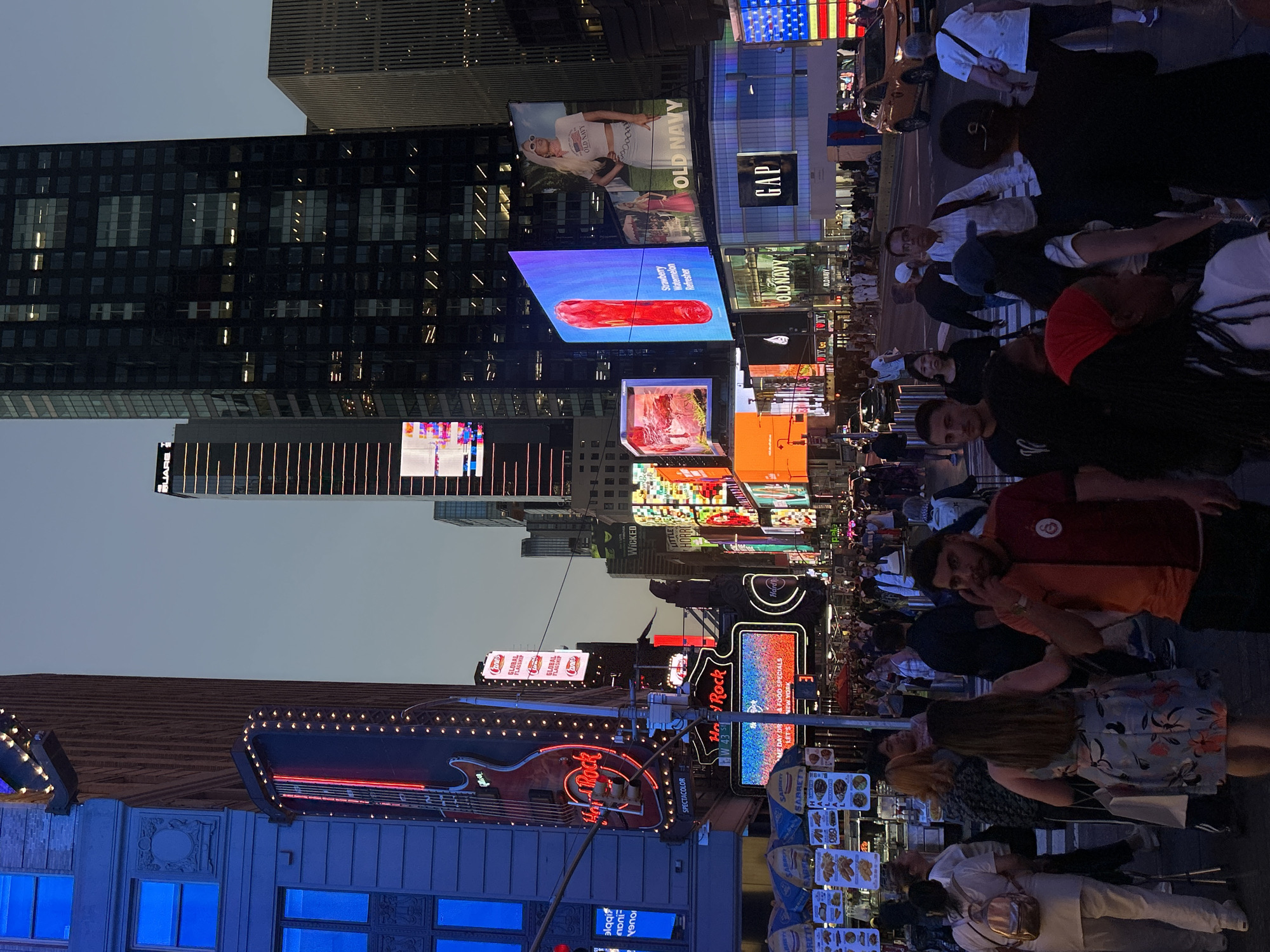 The image captures a vibrant, bustling night scene in Times Square, New York City. The primary subjects are a large crowd of people navigating the busy streets, surrounded by towering buildings adorned with numerous brightly lit digital billboards and neon signs.
The activity taking place is typical of a public space in a major city: people are seen walking, standing, and observing the dazzling advertisements. The atmosphere is energetic and lively, indicative of a popular tourist destination.
Notable details include the time of day, which is clearly night or late dusk, given the dark sky contrasted with the intense artificial light from the signs. The weather appears clear. Location cues such as the iconic architecture, the sheer density of commercial displays, and specific brand names strongly identify the setting as Times Square.
Visible text includes prominent brand names like "Old Navy," "GAP," and "Hard Rock," the latter featuring its iconic neon guitar sign. Other advertising copy visible includes "Strawberry Watermelon Refresher," "Wicked," "Sabrett" (likely a hot dog vendor), and "Canes GLOBAL FLAGSHIP." An American flag display is also visible on one of the screens. The overall impression is one of overwhelming visual stimulation and commercial activity.