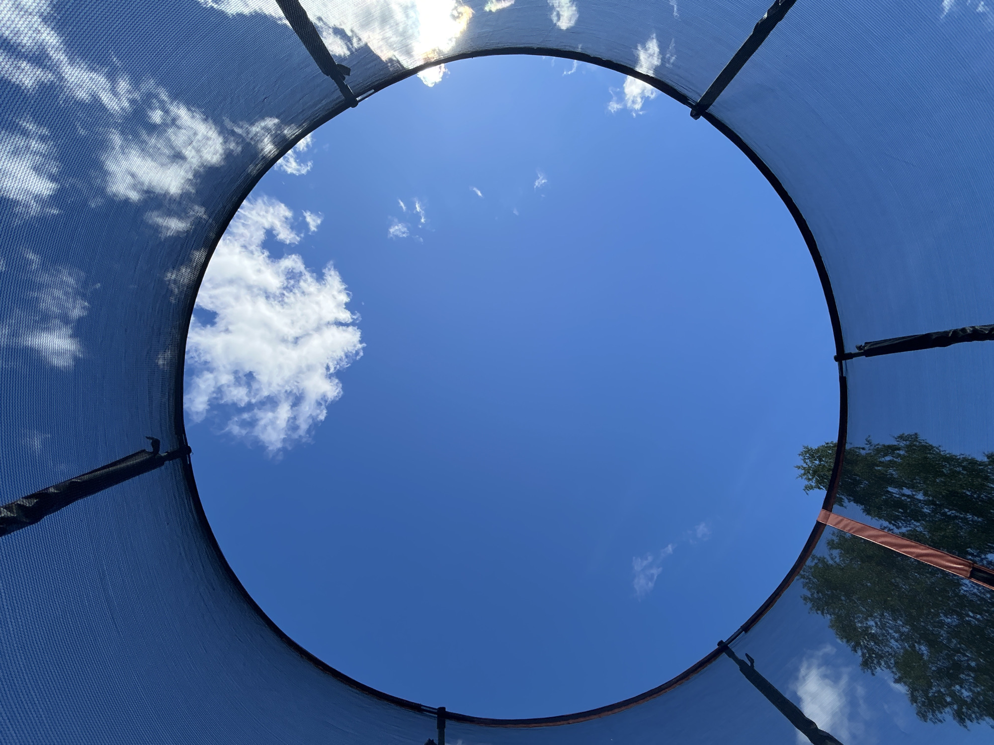 The image shows a view looking up through the netting of a trampoline towards a bright blue sky with scattered white clouds. The edge of the trampoline's protective netting forms a circular frame around the sky. Trees are visible in the lower left portion of the frame, seen through the netting. The scene appears to be outdoors on a sunny day. The location is identified as the city of Novozhilovo, Russia. There are no people or specific actions depicted in the image.