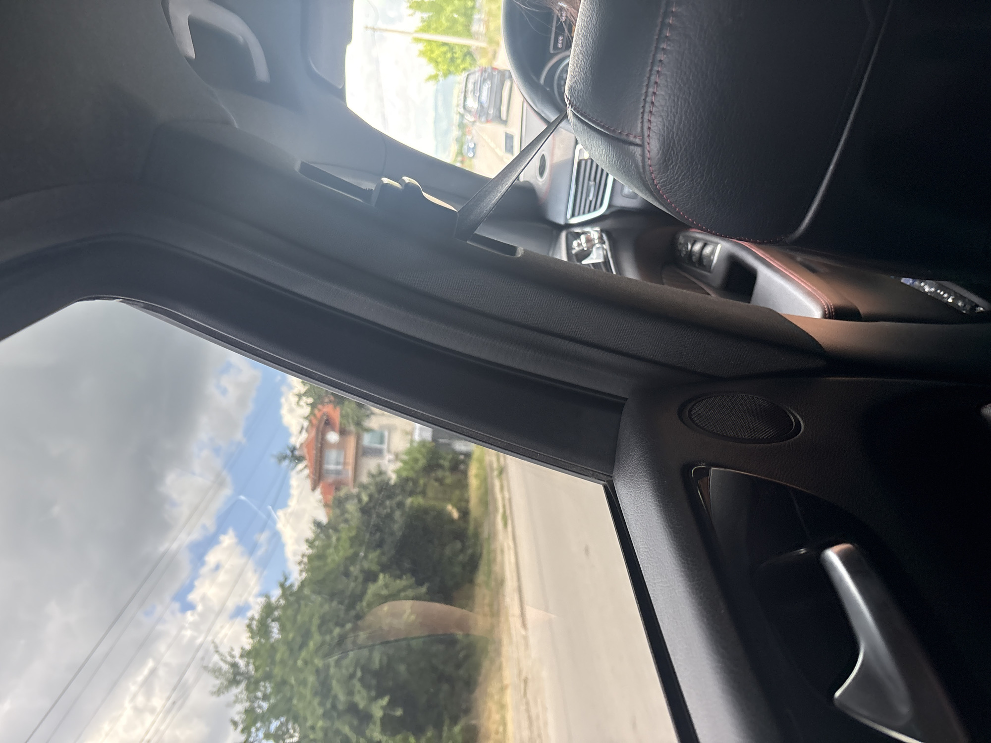 The image is an interior view from a car parked on a street in Elin Pelin, Bulgaria. The perspective is looking out the passenger side window, with the car's interior elements dominating the foreground. The dashboard, a black leather seat with red stitching, and a car door handle are visible. Outside the window, a residential building with an orange roof and lush green trees line a road. The sky is partly cloudy, suggesting it is daytime. The scene appears to be a calm, everyday moment. No people are clearly visible, and no text is present in the image.