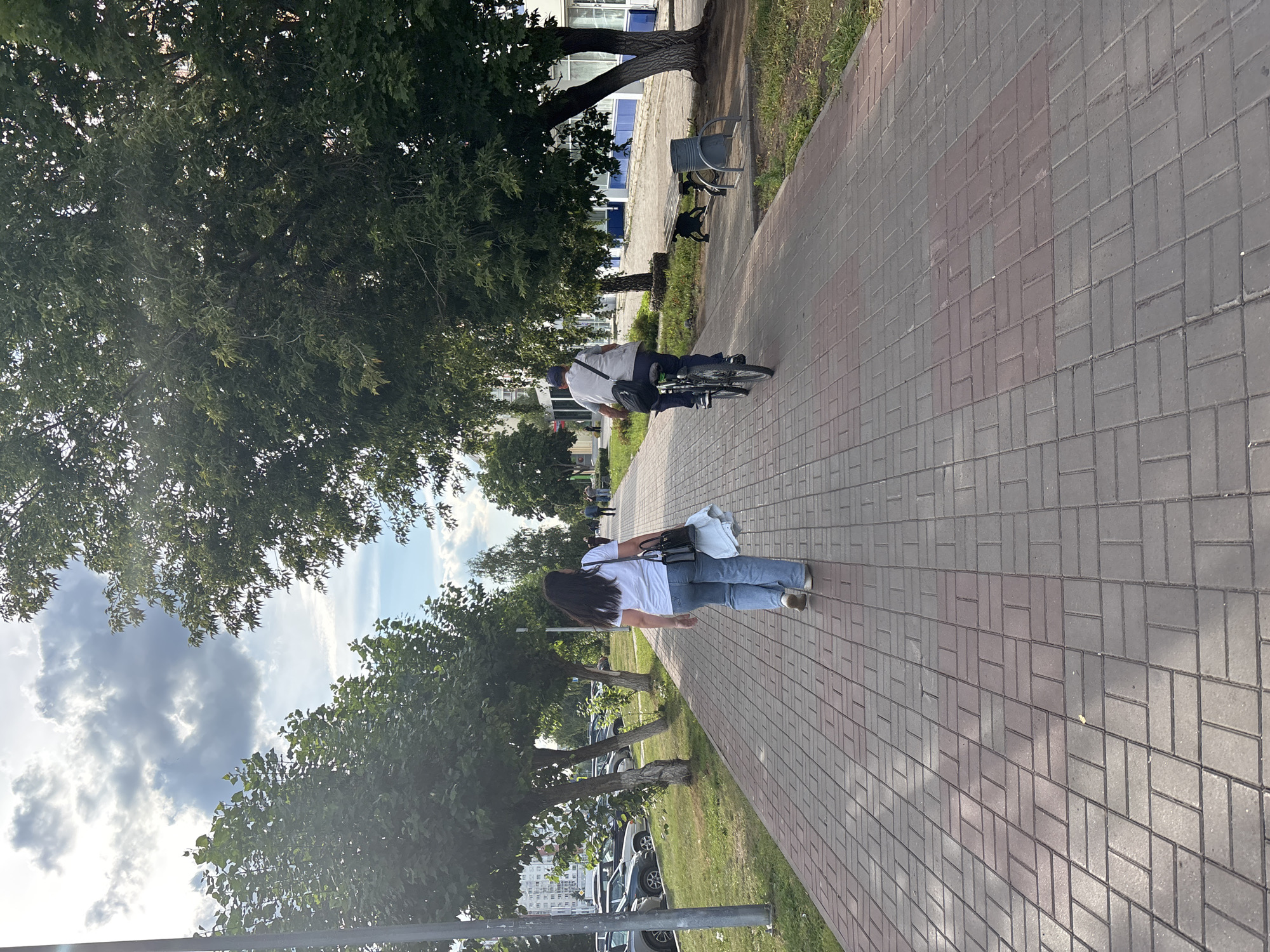 In Naberezhnye Chelny, Russia, a woman walks away from the camera on a brick sidewalk. She wears a white t-shirt and blue jeans, carrying a black purse and a light-colored garment. A man on a bicycle rides away from the camera on the same sidewalk, wearing a light-colored shirt and dark pants, with a black bag slung over his shoulder. The sidewalk is bordered by a grassy area and trees, with parked cars visible to the left and buildings in the background. The sky is partially cloudy.