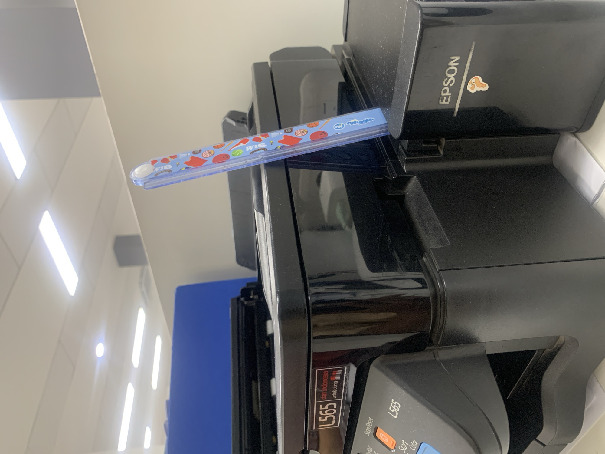 The image shows a close-up of a black Epson L565 printer, with a blue plastic ruler adorned with cartoon candies stuck into its side. The ruler is leaning against the printer's right-hand side, with its top extending upwards. The printer has various buttons and labels, including "Stop/Reset", "Start Color", and "L565". A sticker with the "EPSON" logo is visible on the right side of the printer, with a small cartoon sticker of a shrimp below it. The background shows a sterile office environment with fluorescent lighting and a blue partition. The image does not provide any specific details about the city of Palembang, Indonesia, other than the text on the printer which reads "dan Indonesia" and "untuk dunia".