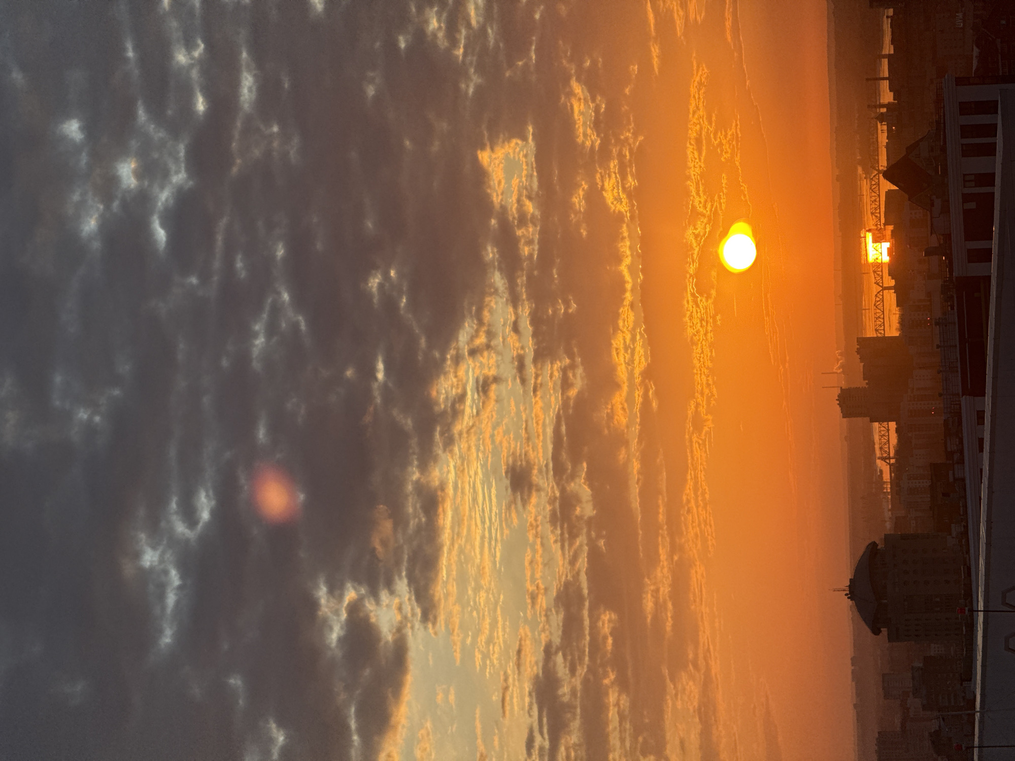 The image captures a sunset in Perm, Russia, presenting a bright orange sun near the horizon. To the right of the sun, there's a bridge partially visible, possibly the Kama Bridge, amidst a cityscape. Below the bridge, buildings form the skyline, their details somewhat obscured by the atmospheric haze. A prominent building with a distinctive dome-like structure is situated near the bottom of the frame. The sky is filled with clouds, ranging from dark grey to lighter shades of orange, reflecting the setting sun. The overall composition suggests an urban landscape bathed in the warm hues of the setting sun.