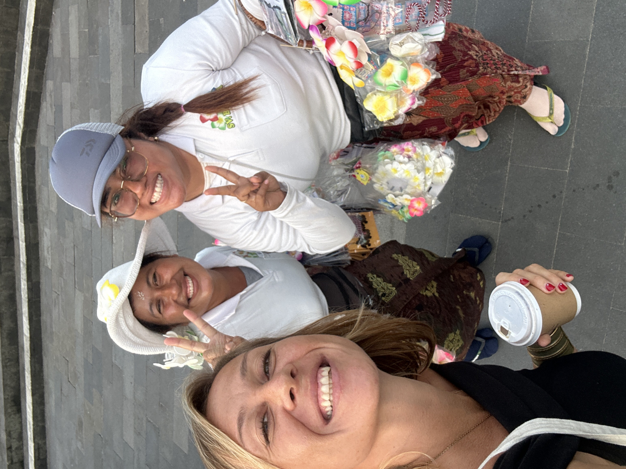 A selfie taken outdoors shows three women. The woman closest to the camera, on the left, has blonde hair and is smiling. She is wearing a black top and holding a cup of coffee in her right hand, which has red nail polish. Two women are behind her. The woman in the center is wearing a white hat with a yellow flower and a white shirt, and she is smiling and holding up two fingers in a peace sign. She is wearing a patterned sarong. The woman on the right is wearing a gray baseball cap, glasses, and a white shirt. She is also smiling and holding up two fingers in a peace sign. She is carrying a basket of colorful flower ornaments. The background appears to be a stone pathway or steps. The presence of traditional Indonesian clothing and the style of the flower ornaments suggest the location may be in Indonesia, but there is no definitive information to confirm it is in the city of Kediri, Indonesia.
