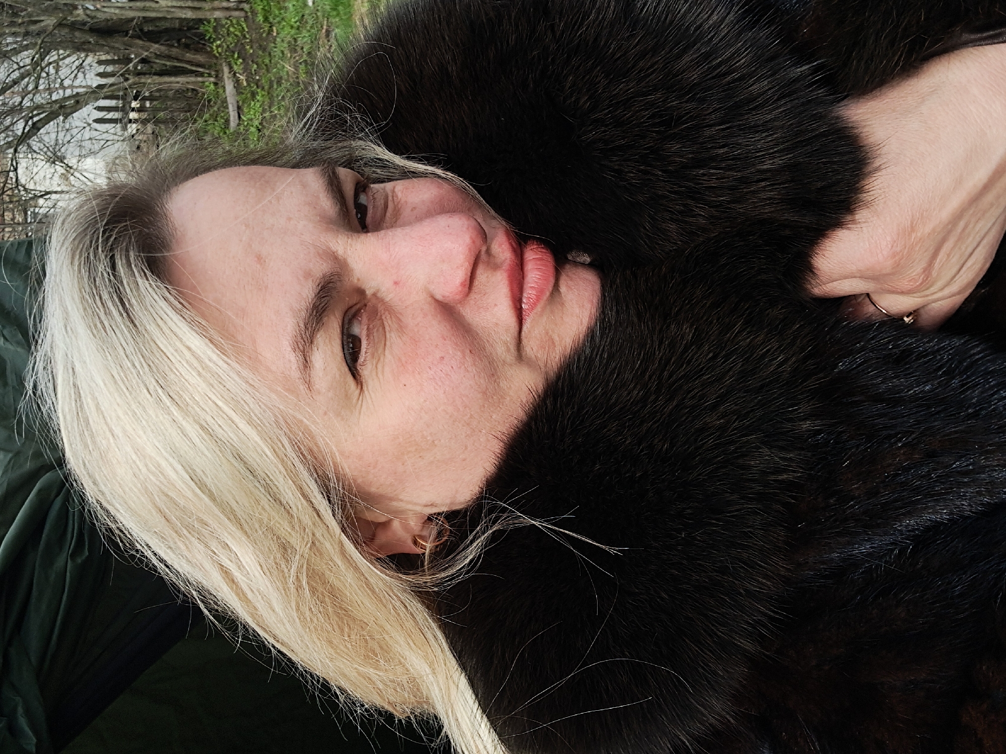 A woman with blonde hair, wearing a dark fur garment, is the central subject. She appears to be lying down, and the background includes sparse trees and vegetation, possibly near a wooden fence. The location is not explicitly discernible from the image. There is no information on the location of city Полянки, Russia. The woman is wearing a ring on her right hand.