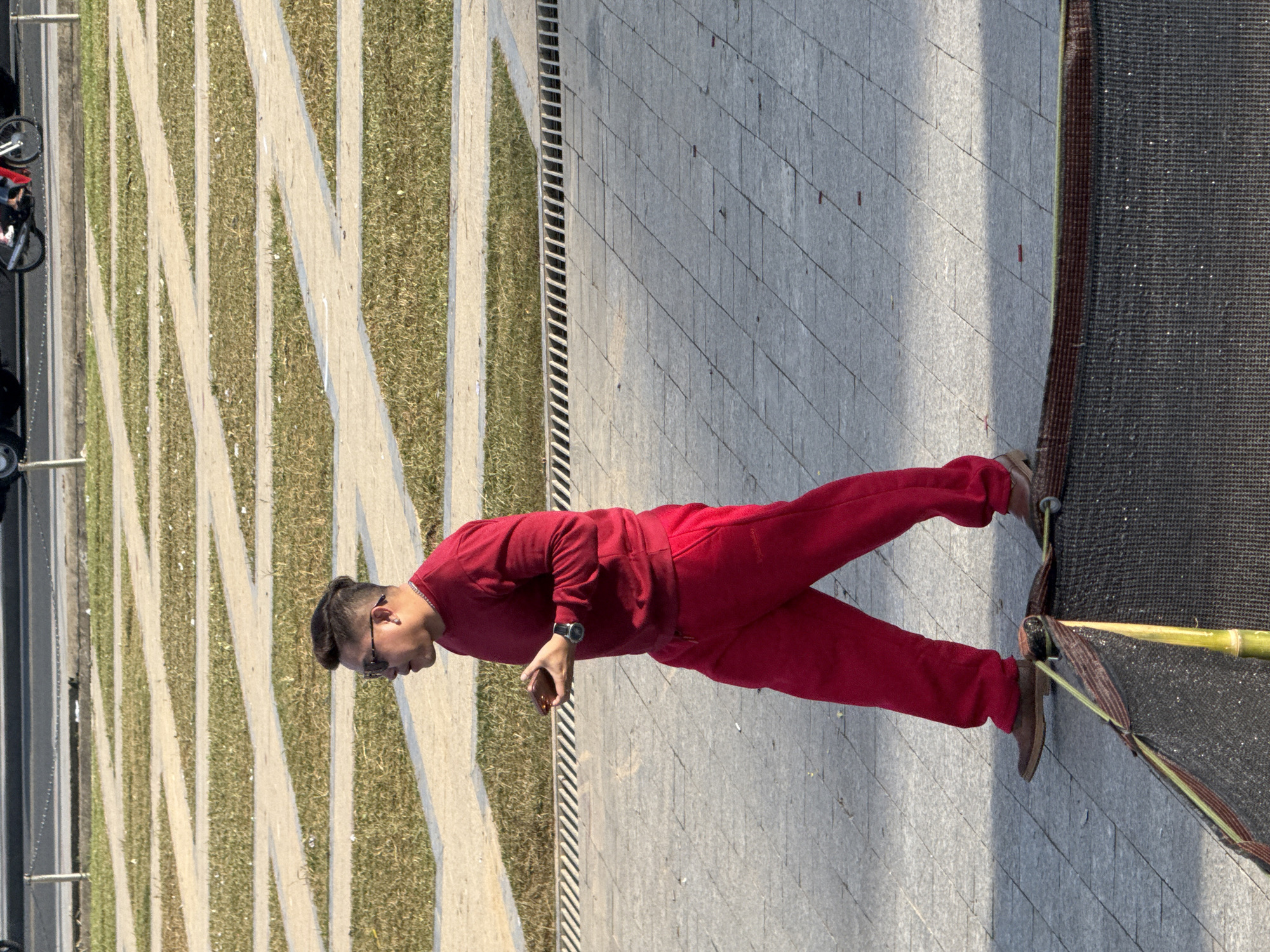 A full-body, side-view shot captures a Vietnamese man walking on a paved area in Da Lat, Vietnam. He is dressed in a red long-sleeved shirt and matching red sweatpants. The man is wearing sunglasses and a watch on his left wrist. In his right hand, he holds a brown rectangular object, possibly a phone or wallet. The background features geometric patterns of grass and concrete strips, characteristic of modern urban landscaping. A metal grate runs along the edge of the paved area, separating it from the grassy section. In the distance, a motorcycle with riders is visible on a road, suggesting a city setting. The lighting indicates it is daytime, with shadows cast on the ground. A section of dark netting with a bamboo pole is in the foreground, partially obscuring the lower portion of the man's legs.