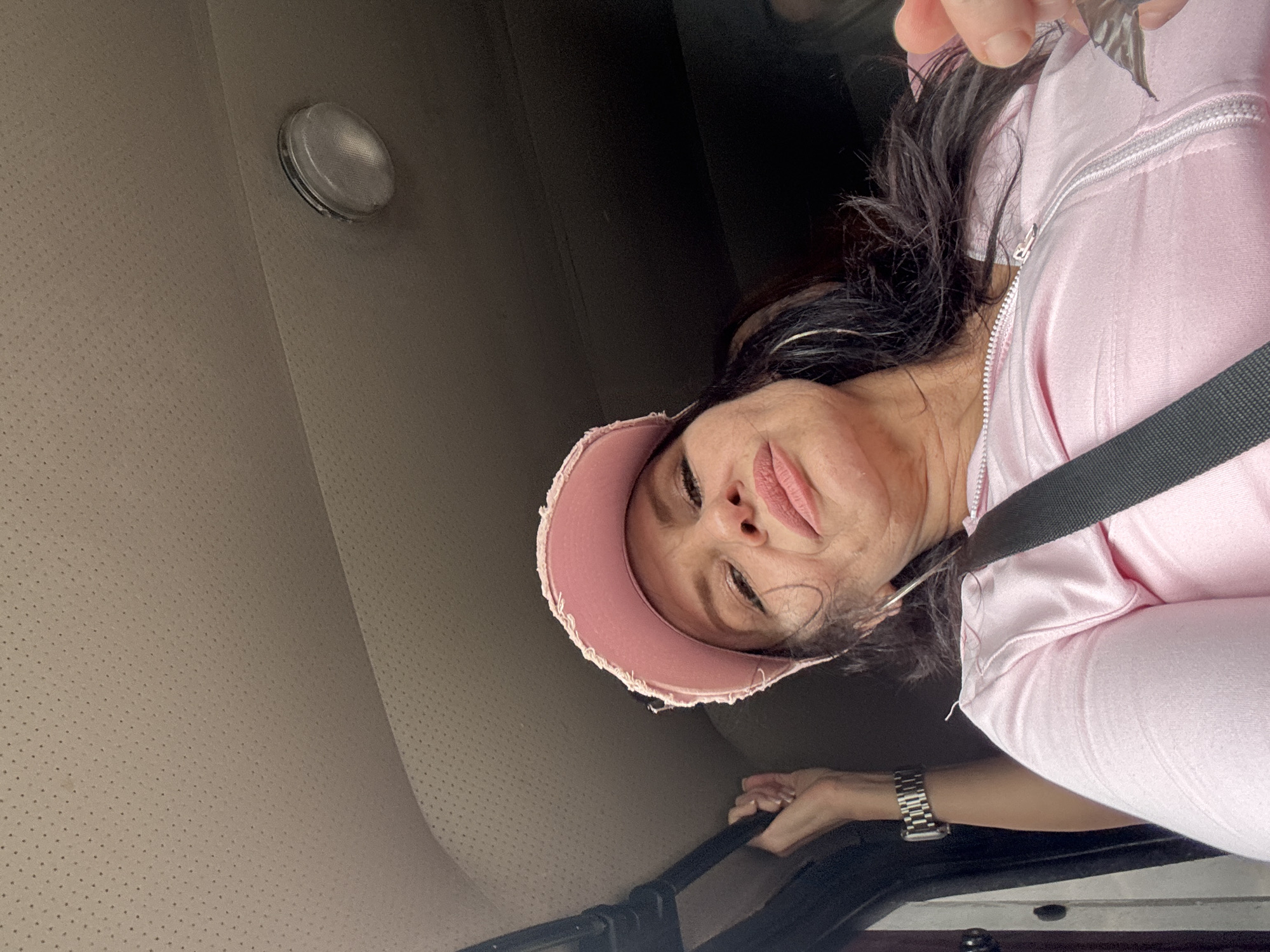 A woman in a pink baseball cap and pink zip-up jacket is in the passenger seat of a vehicle. She is looking down at the camera with her mouth slightly open. A hand is visible in the upper left corner holding onto a handle. The vehicle's interior, including the headliner and a light fixture, is visible. The scene is not identifiable as being in Каменномостское сельское поселение, Russia.