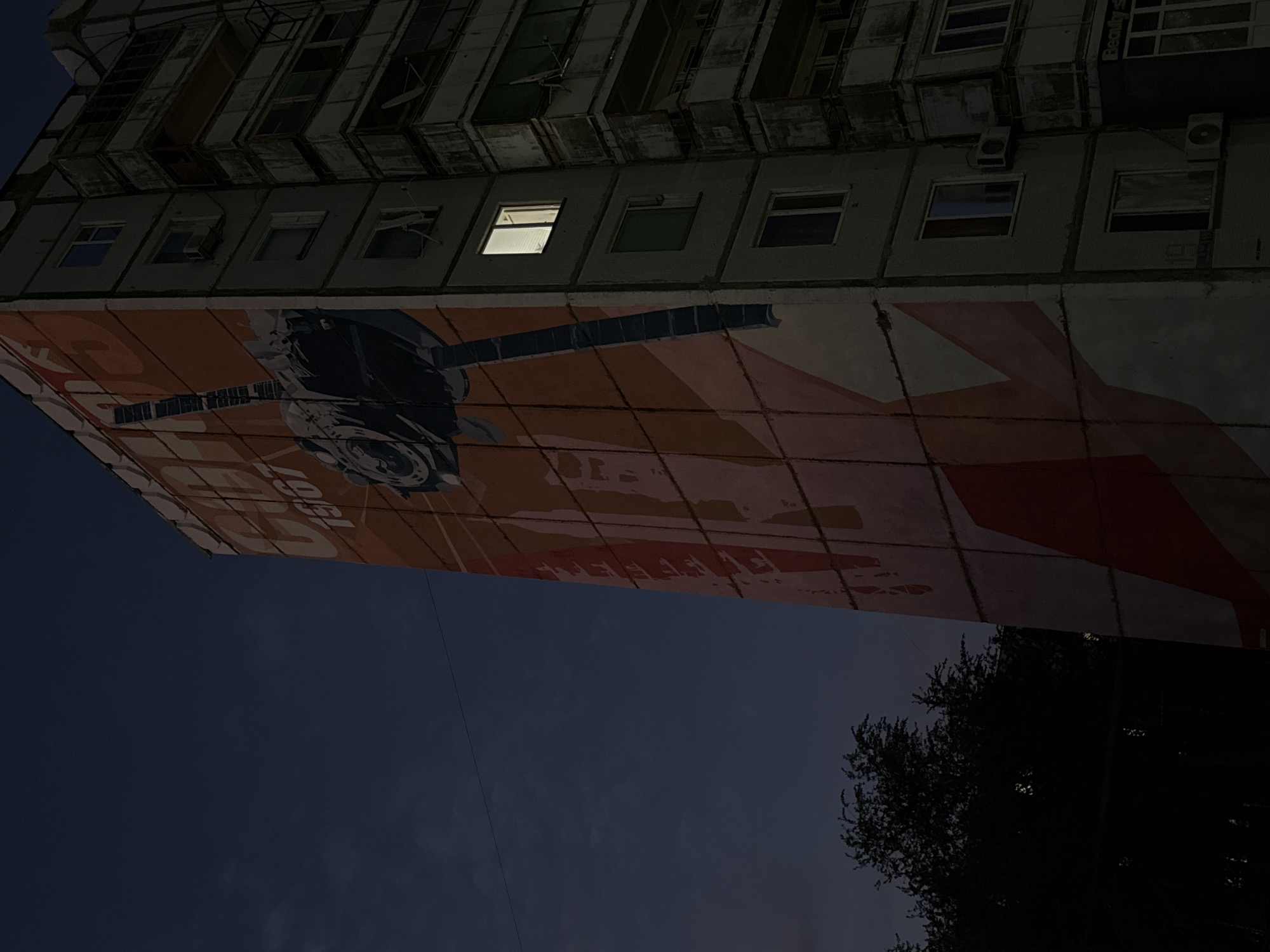 A tall, multi-story residential building is depicted at dusk in Baikonur, Kazakhstan. The building's facade is partially covered by a large, faded mural featuring a stylized image of a satellite and the year "1967." Several windows on the building are visible, with one on the right side illuminated. The sky is a deep blue, transitioning from twilight, and dark trees are silhouetted on the left side of the frame. The overall impression is one of a quiet evening in a Soviet-era urban environment.
