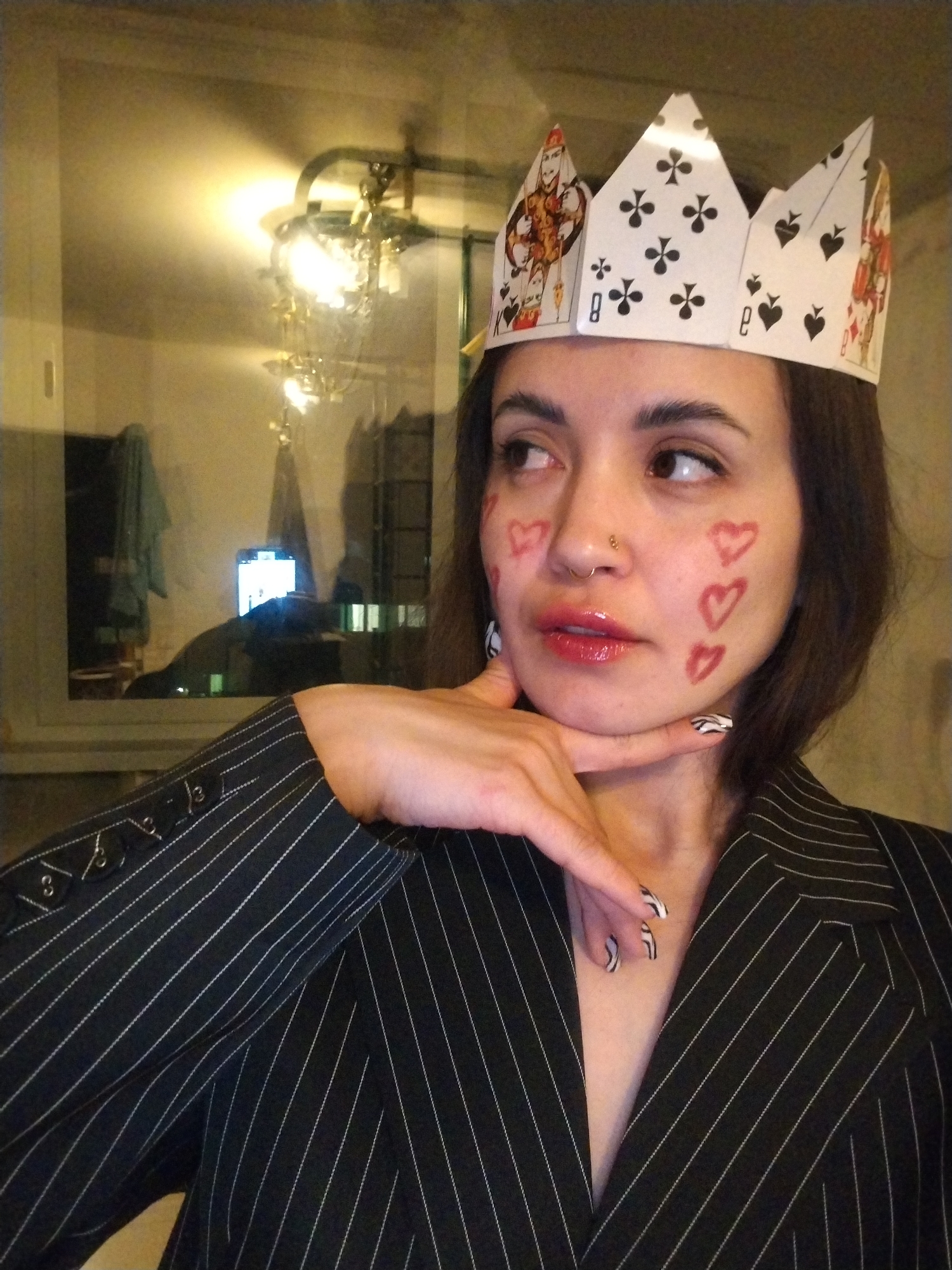 This medium shot depicts a young adult female, likely in her 20s, posing indoors in Noviy, Russia. She is adorned with a creative homemade crown fashioned from playing cards, prominently featuring the King and Queen of Hearts, King of Clubs, and cards displaying the 8 of Clubs and 8 of Spades. Her face shows glossy red lipstick and small red heart shapes drawn on her cheeks, complemented by a subtle nose piercing.
She wears a dark pinstripe blazer with thin white stripes, and her left hand, with nails painted in a distinctive black and white zebra pattern, rests thoughtfully under her chin. Her gaze is directed to her right (viewer's left), suggesting a pensive or contemplative mood.
The background reveals a dimly lit indoor setting. Reflections in what appears to be a window show an illuminated chandelier or light fixture, and the bright windows of an outside building, indicating it is likely evening or nighttime. The playful costume elements suggest a themed event, perhaps a "Queen of Hearts" motif or a festive occasion. Aside from the playing card markings, no other significant text is discernible.