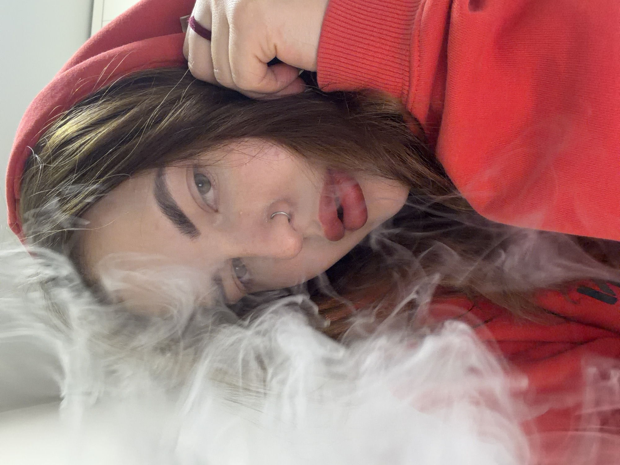 An indoor portrait features a young individual, likely female, with long brown hair, wearing a red hooded sweatshirt. The image is rotated counter-clockwise, portraying the subject from a low-angle perspective, possibly lying down or leaning back, with her gaze directed towards the camera. A significant cloud of white smoke or vapor billows from her mouth, obscuring part of her face and extending into the foreground, suggesting vaping or smoking. Visible facial features include one eye, a silver nose ring on her left nostril, and prominent red lips. One hand is visible near the top of her head. The background is a plain, light-colored surface. The setting is indoors in Krasnodar, Russia.