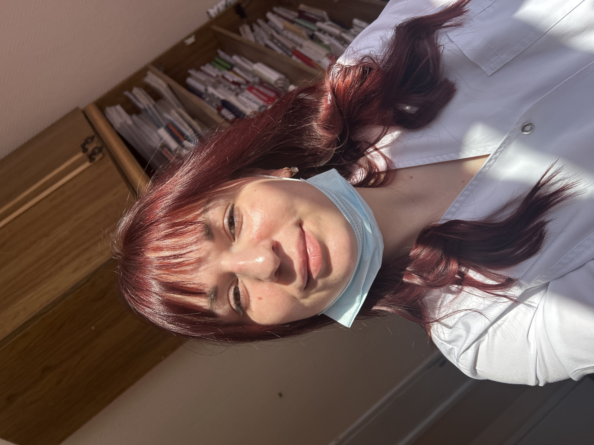 A young woman with auburn hair is wearing a light blue surgical mask and a white lab coat. She is indoors. In the background is a wooden cabinet filled with papers and files. The light suggests it's daytime. It is not possible to know the city.