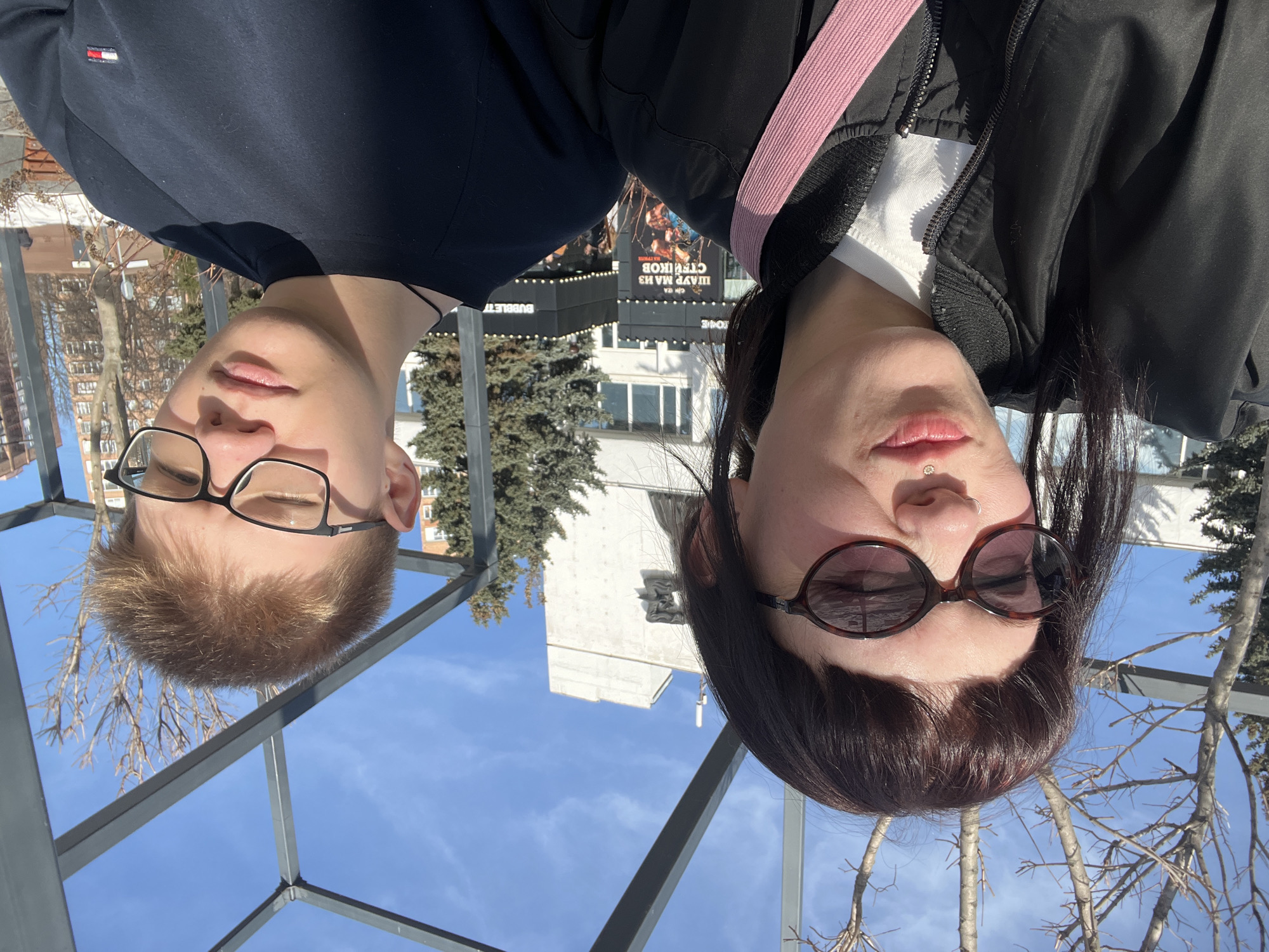 A selfie-style image depicts two young adults, one female and one male, outdoors under bright sunlight against an urban background in Izhevsk, Russia, likely near K. Marksa Street, 246A.
The female, positioned on the left, has dark hair with bangs, wears oval dark sunglasses, a silver nose ring, and a silver labret piercing (medusa) with a small gem. She is wearing a black bomber jacket over a white top, with a pink fabric strap visible across her chest. Her lips are slightly puckered, and she looks towards the camera.
The male, positioned on the right, has short, light brown hair and wears black-framed glasses; his eyes are mostly closed or squinting due to the sun. He wears a dark blue long-sleeved shirt with a small red, white, and blue logo on the left chest, and a thin black cord is visible around his neck.
The background features a clear blue sky with some wispy clouds. Prominent dark grey metal structural beams, possibly part of a pergola, frame the top of the image. Visible beyond the individuals are a large, light-colored building with a distinctive upper section and a potential antenna, along with dark decorative relief. A black storefront with white lettering displays "ШАУР МА ИЗ СТЯКОВ" (Shawarma from Steaks) and "BUBBLETEA." Bare deciduous trees and evergreen coniferous trees are present, alongside patches of snow on the ground, suggesting a late winter or early spring setting. Further in the distance, a multi-story residential building with an orange hue is partially visible.